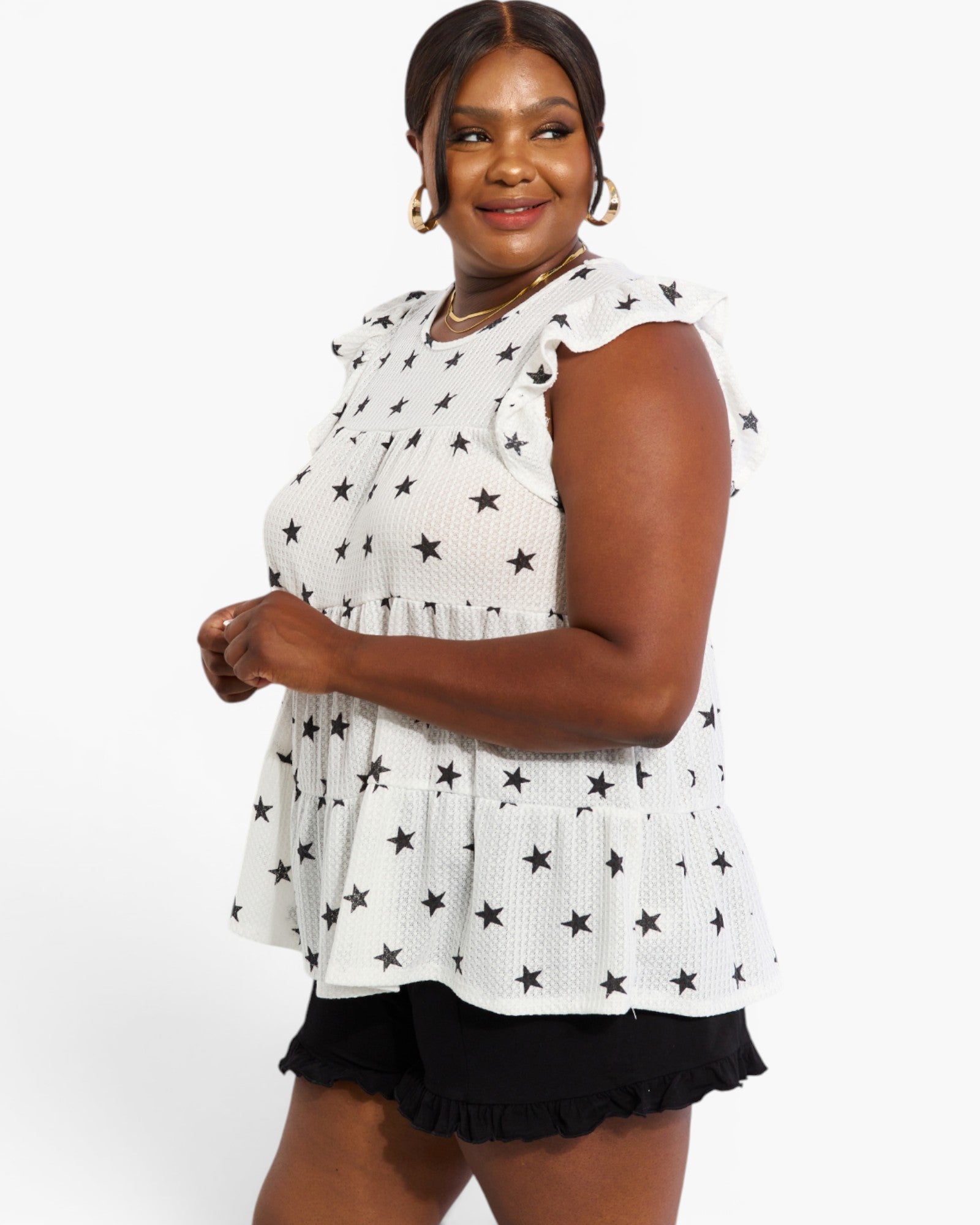 A woman modeling a white sleeveless peplum top with a black star pattern, paired with black ruffled shorts, against a plain white studio background.