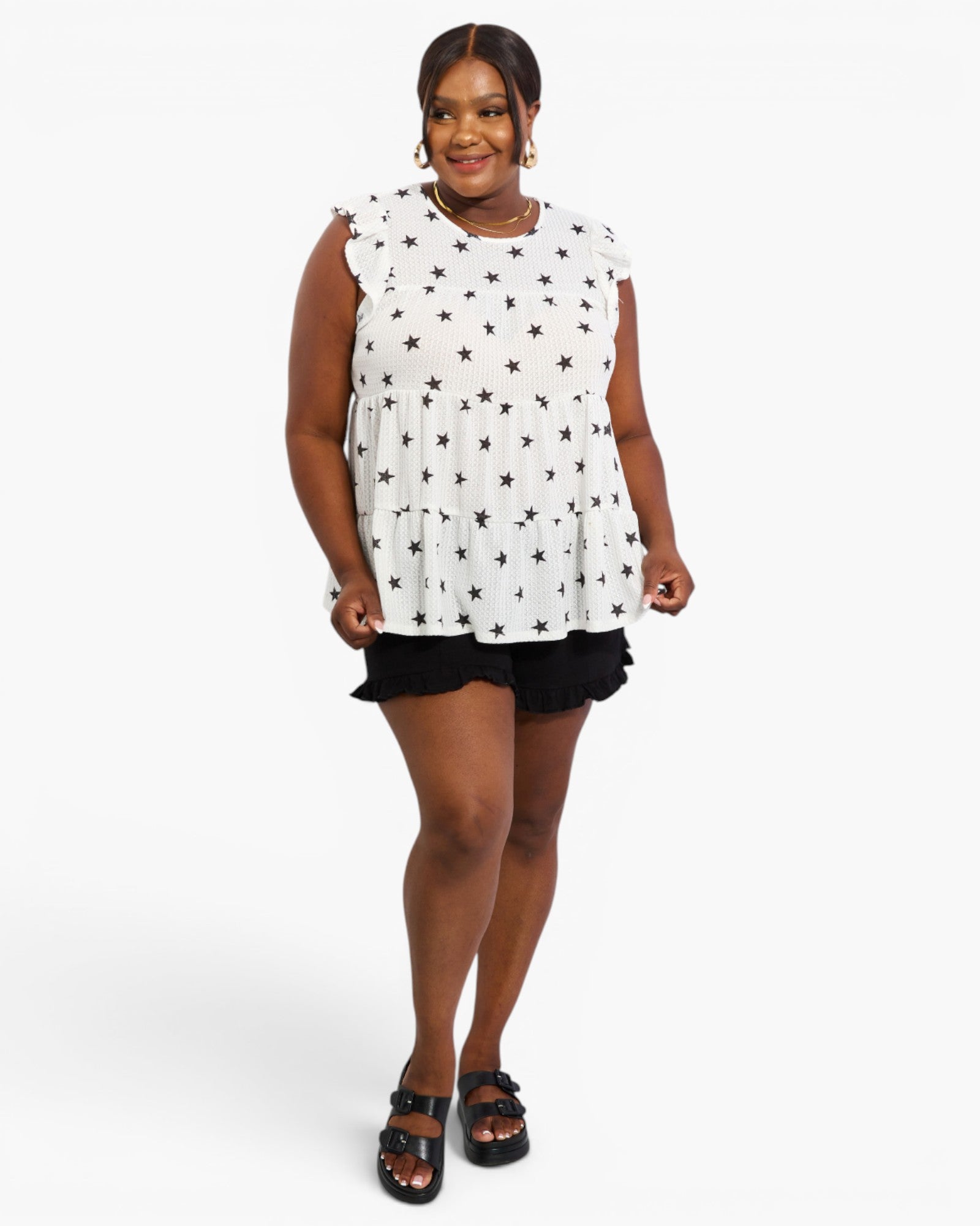 A woman modeling a sleeveless white top with a black star pattern, paired with black shorts and black sandals, against a light gray studio background.
