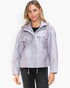 A woman modeling a light lavender hooded jacket with a front zipper, snap-button chest flap pockets, and drawstring hem, paired with a black top and dark jeans against a plain white studio background.
