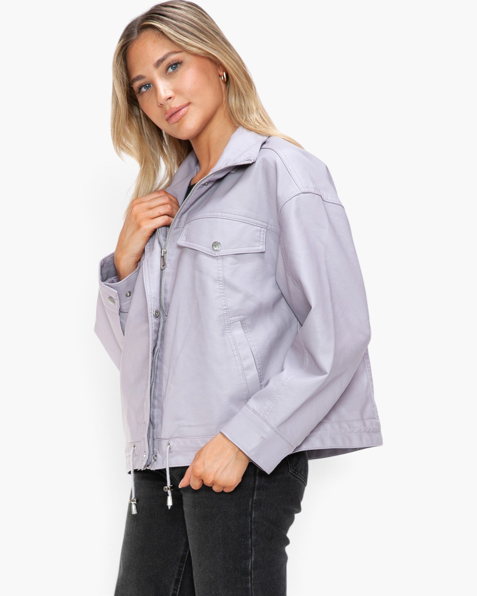 A woman modeling a light lavender-gray utility jacket with a front zipper, buttoned chest pocket, and drawstring details, paired with black jeans against a plain white studio background.