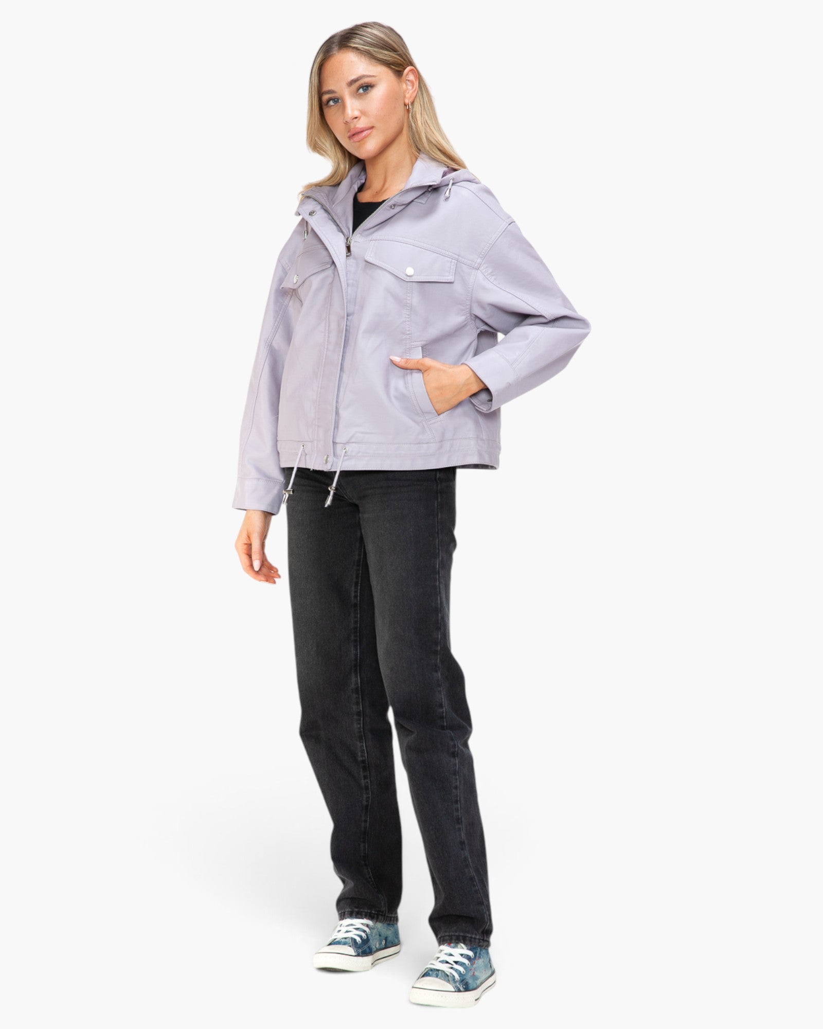 A woman modeling a light lavender-gray cropped utility jacket with a front zipper, chest flap pockets, and a drawstring waist, paired with dark jeans and blue sneakers against a plain white studio background.