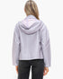 A woman viewed from the back wearing a light lavender hooded jacket and dark gray jeans against a plain white studio background.
