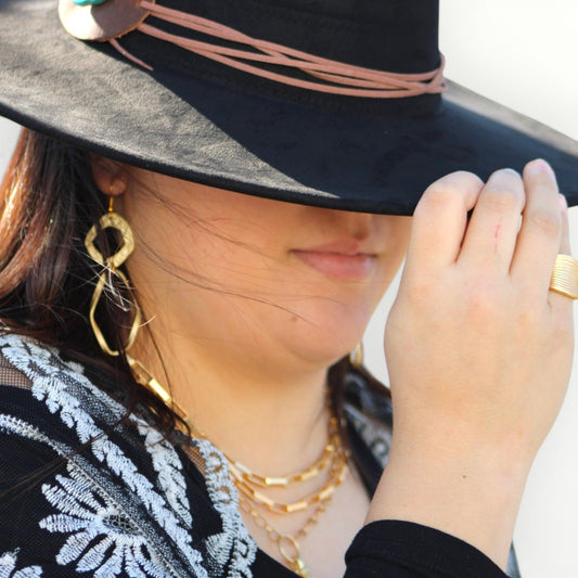 Person wearing a black hat with a pink band, gold earrings, and multiple gold necklaces.