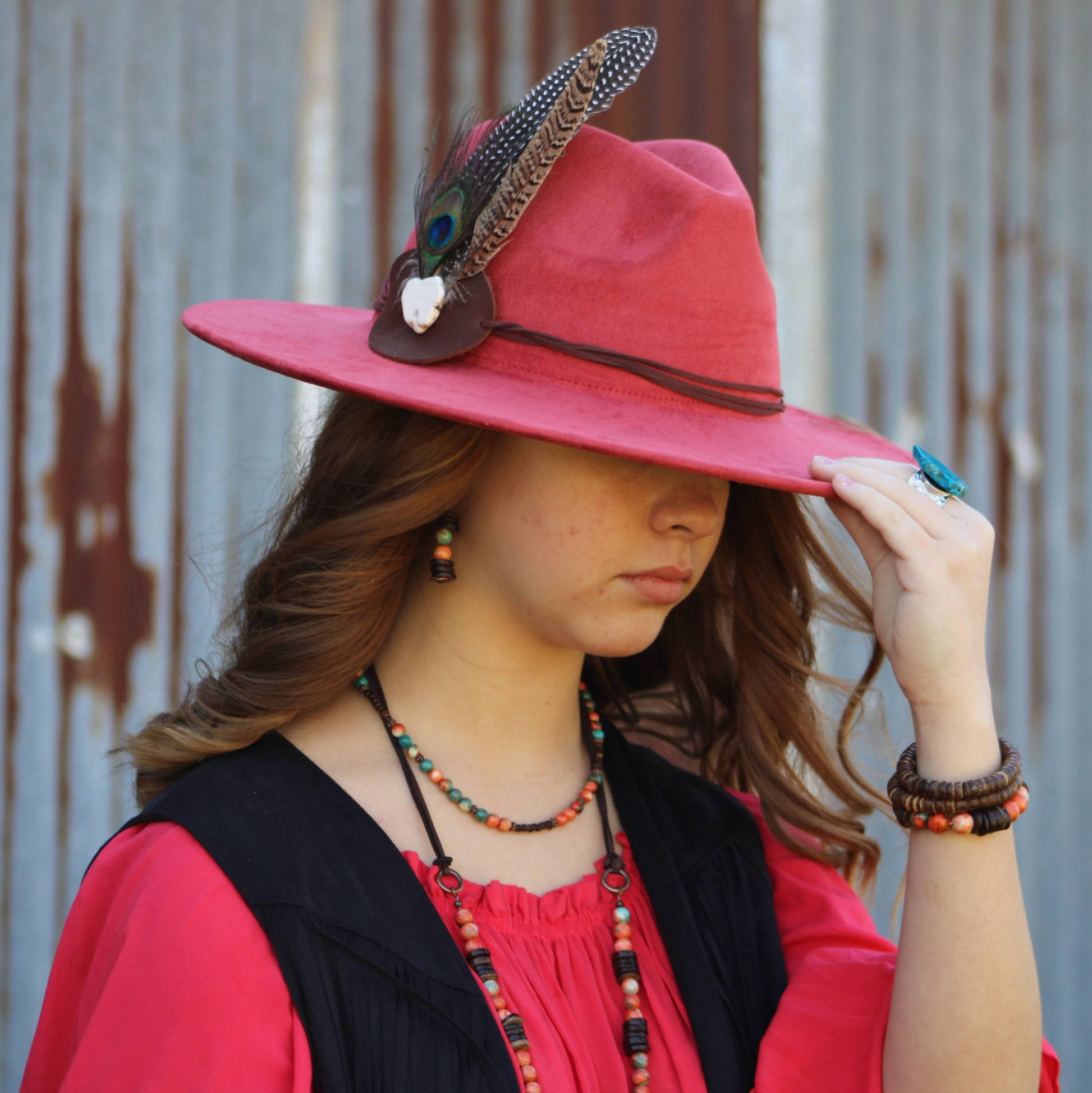 Woman wearing a red hat with feather accents against a rusty metal wall.