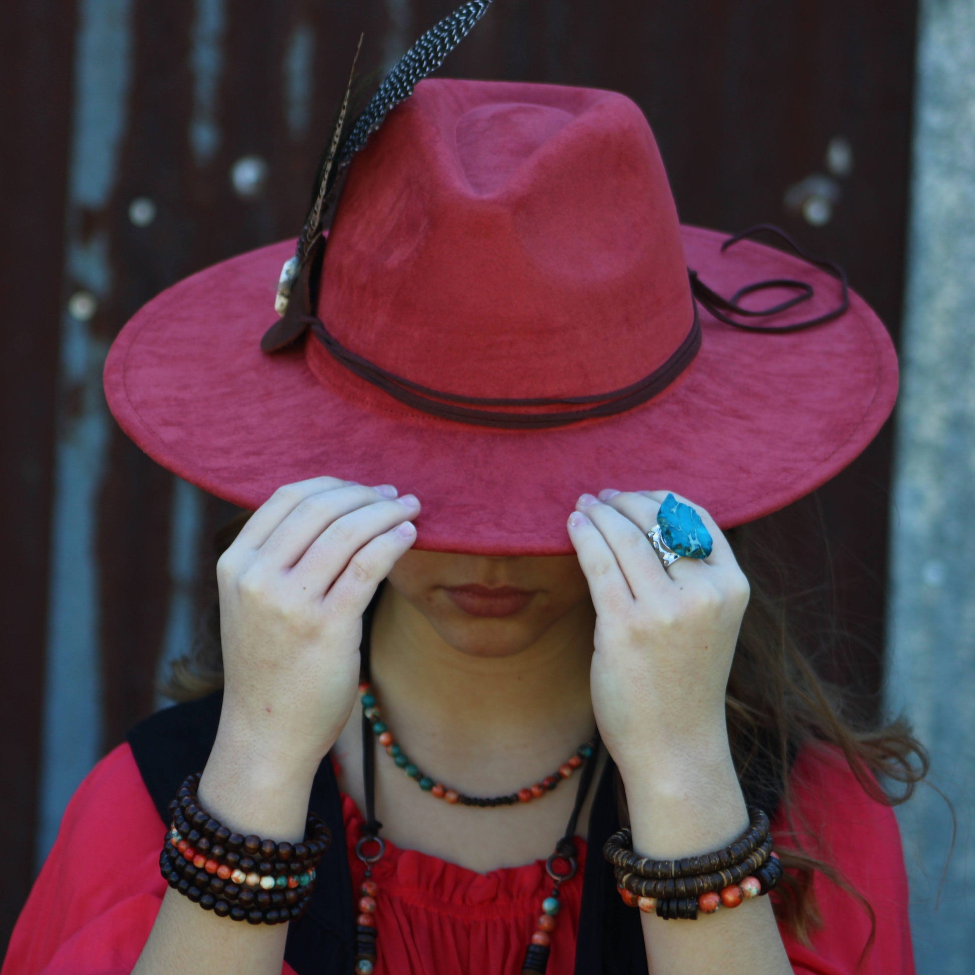 Person wearing a red hat with a feather, holding it over their face, against a blurred background.