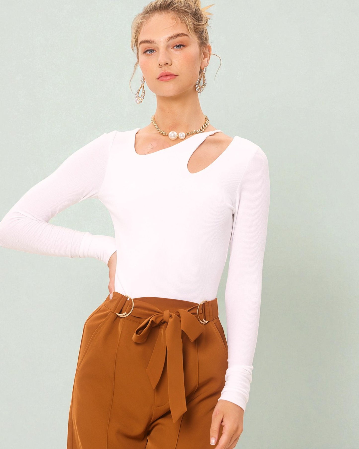Woman wearing a white long-sleeve top and brown pants against a light green background