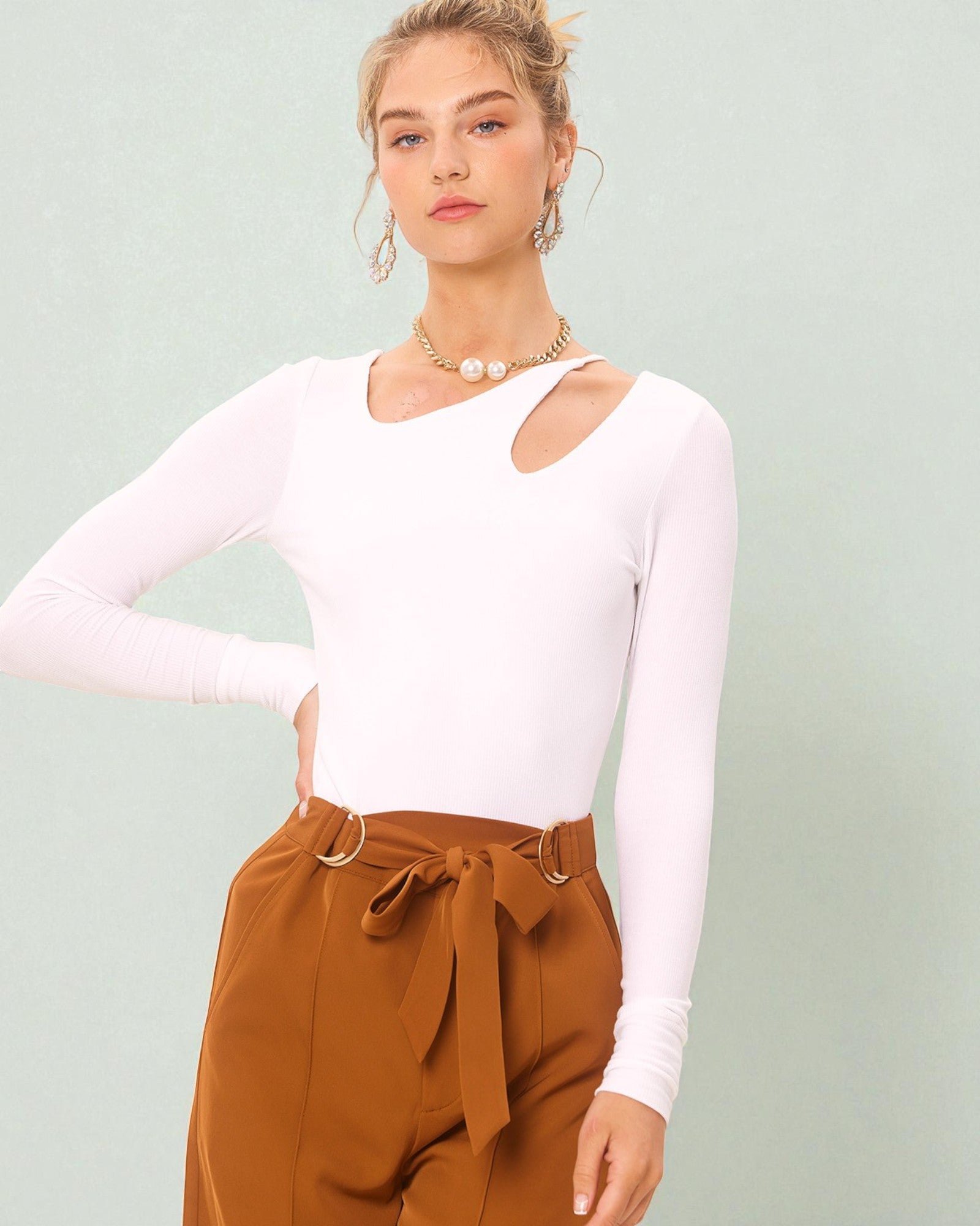 Woman wearing a white long-sleeve top and brown pants against a light green background