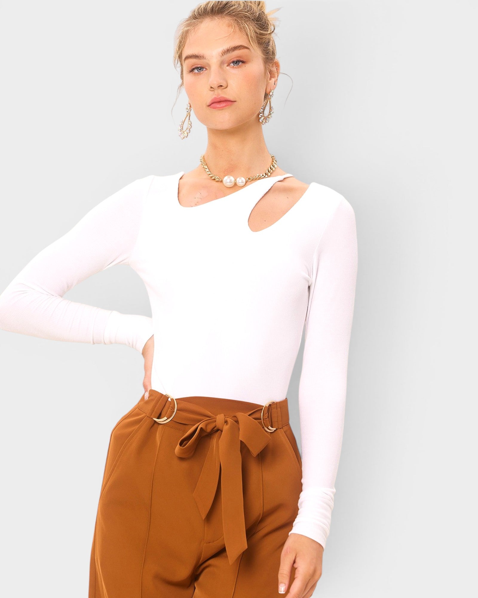 Woman wearing a white long-sleeve top and brown pants with a belt against a light gray background