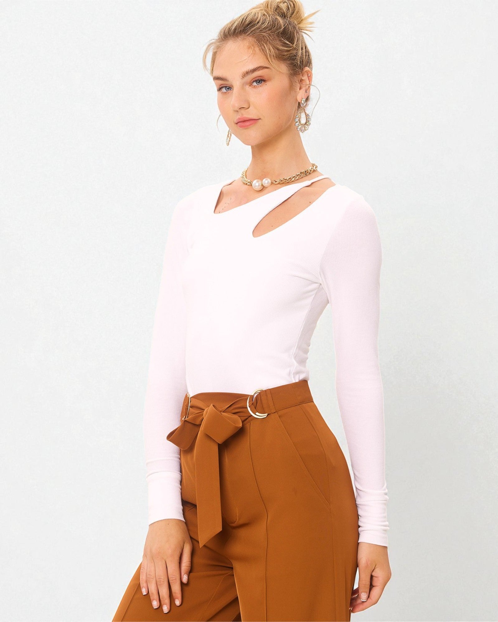 Woman wearing a white long-sleeve top and brown paperbag pants on a light gray background