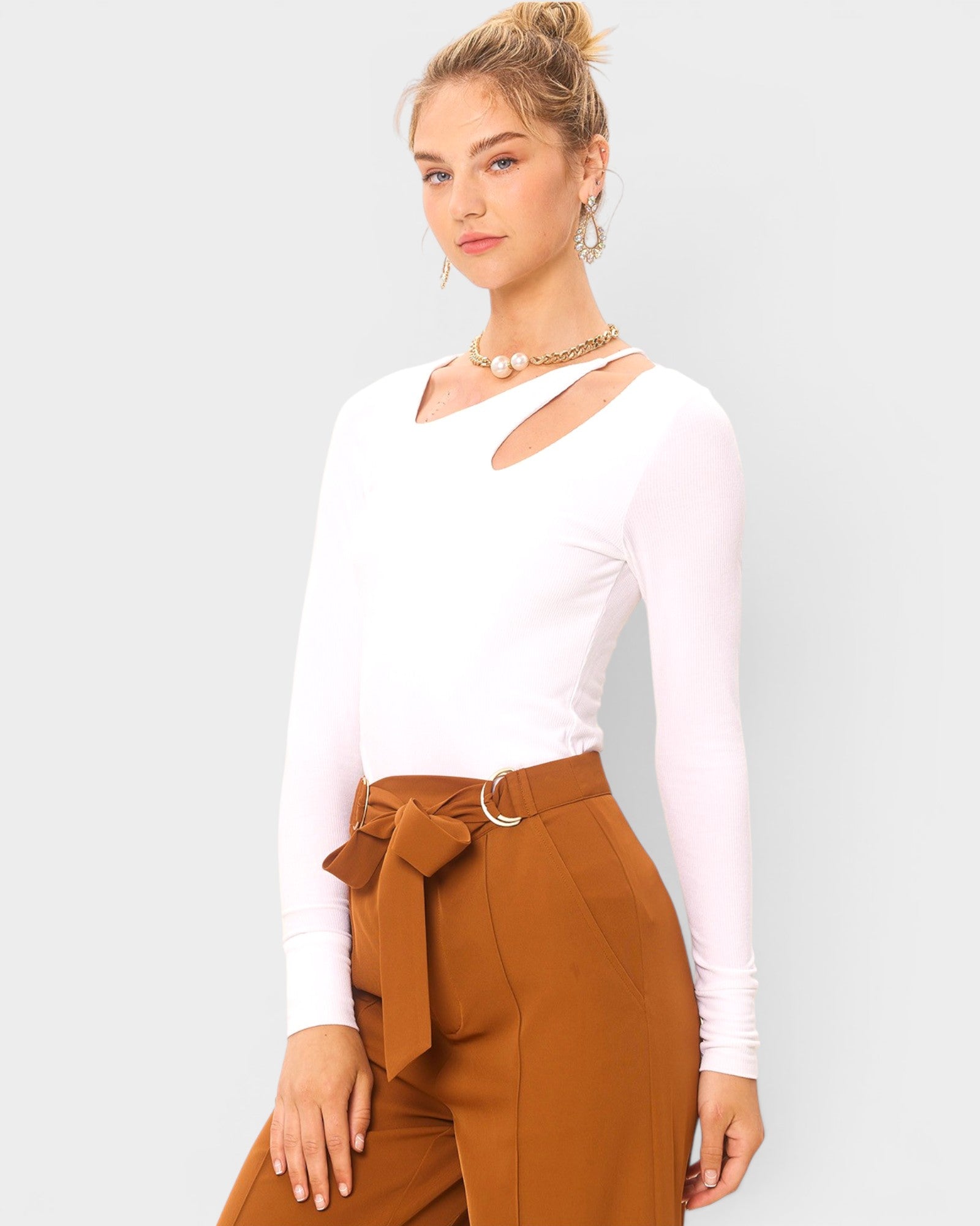 Woman wearing a white long-sleeve top and brown pants with a belt against a plain background