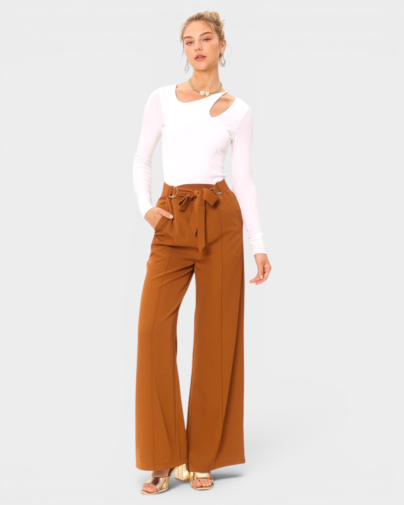 Woman wearing a white long-sleeve top and brown wide-leg pants on a light gray background