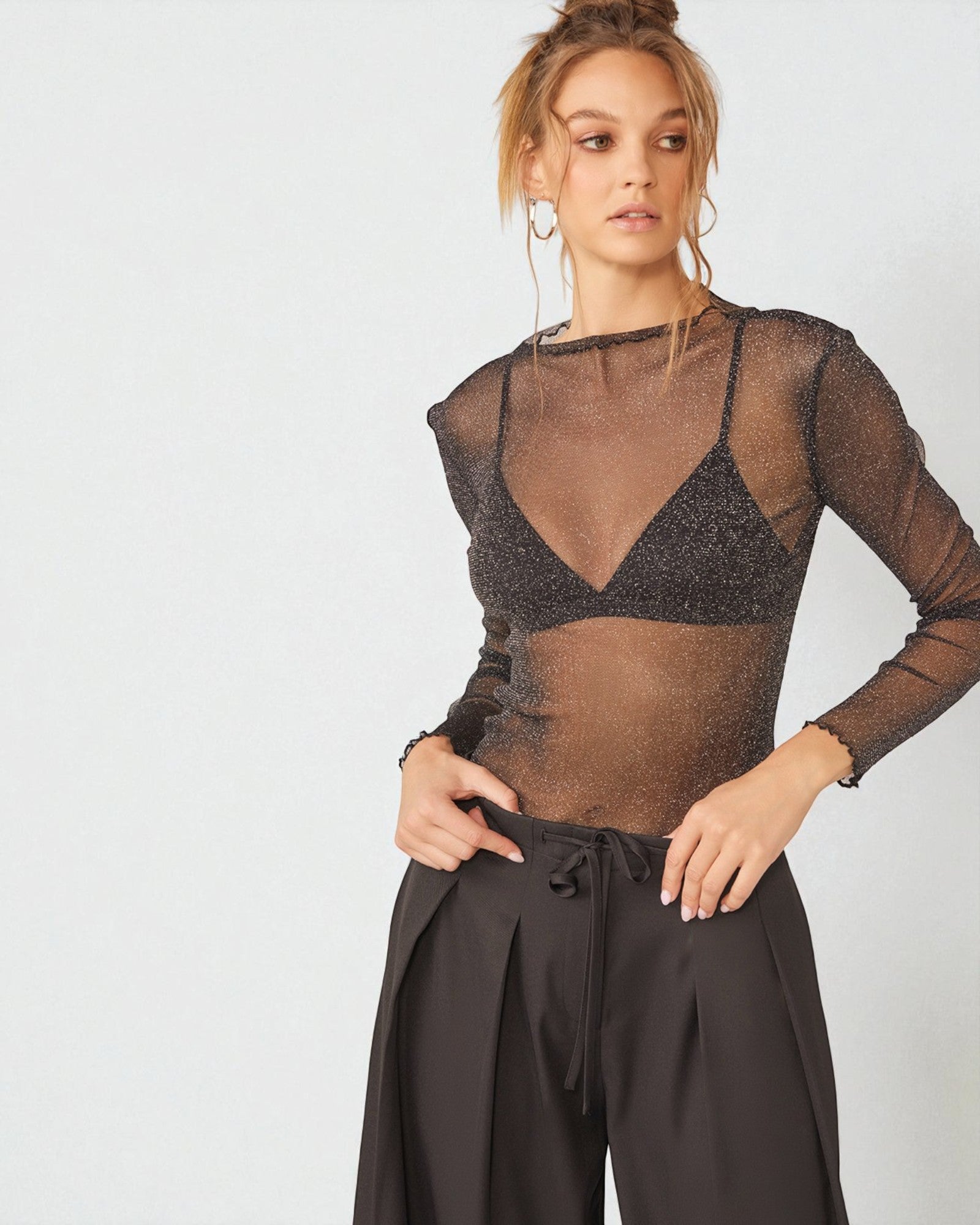 Woman wearing a sheer brown top with long sleeves and high-waisted black pants on a light gray background.