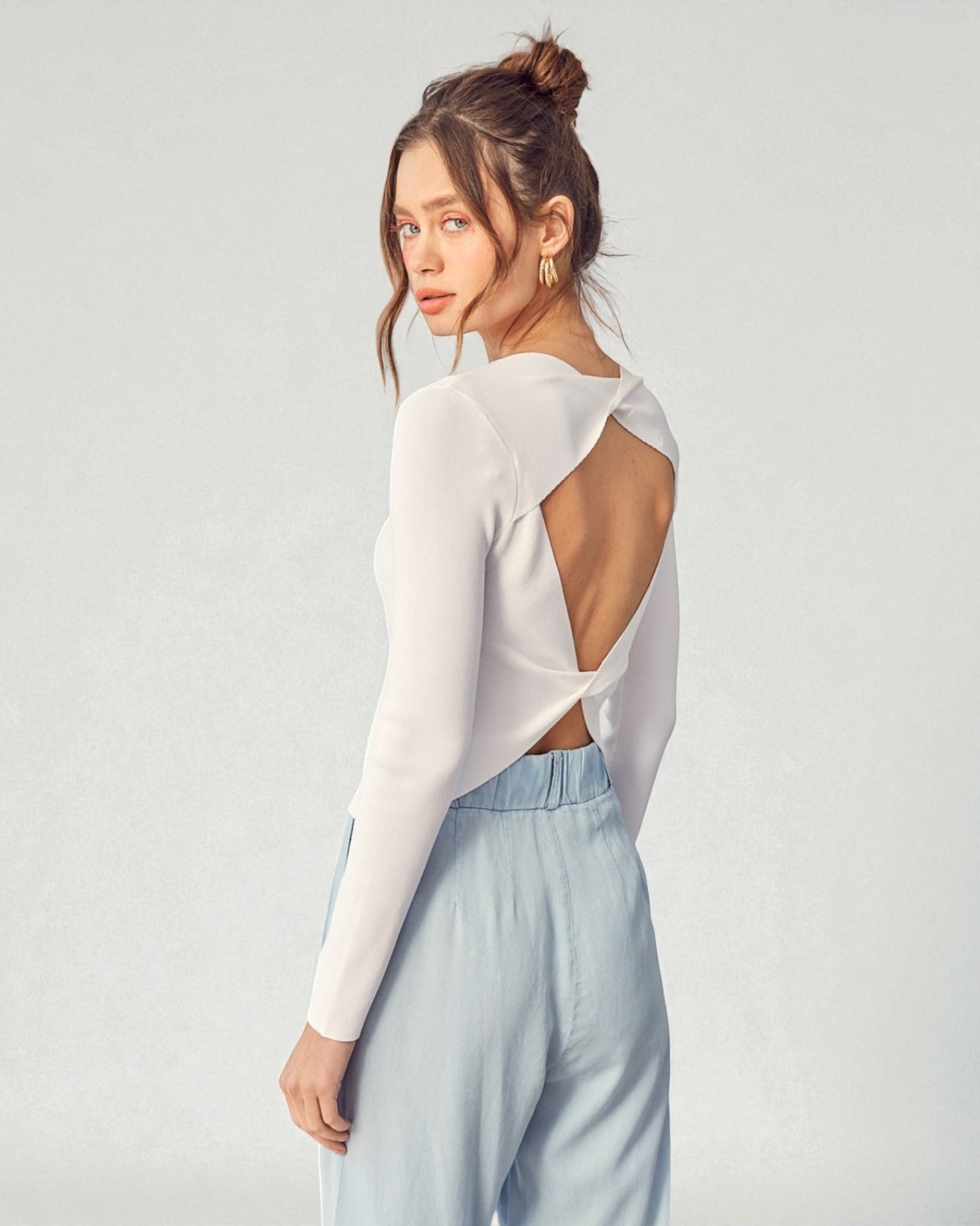 Woman wearing a white long-sleeve top with cut-out design and light blue pants on a plain background