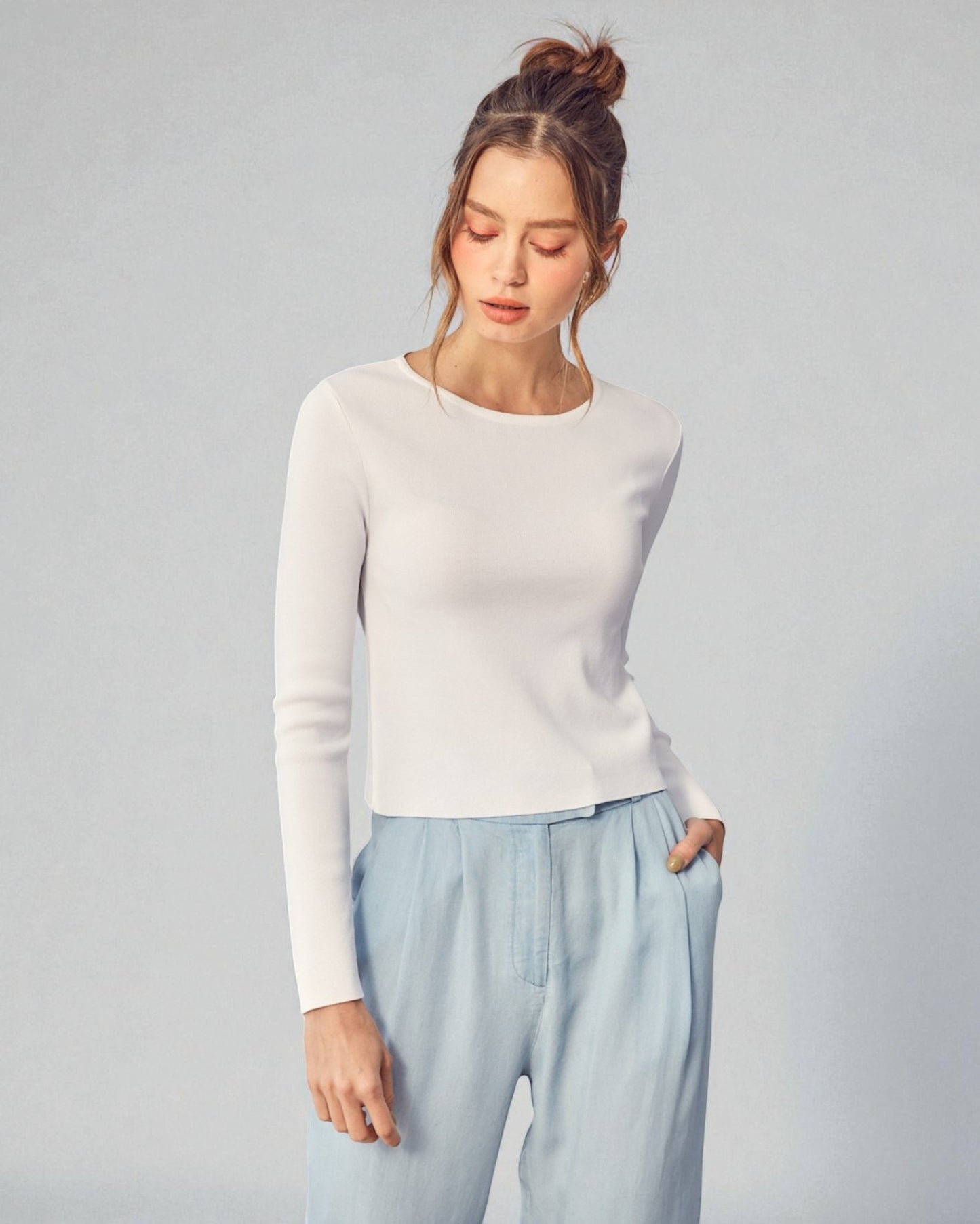 Woman wearing a white sweater and light blue pants against a gray background