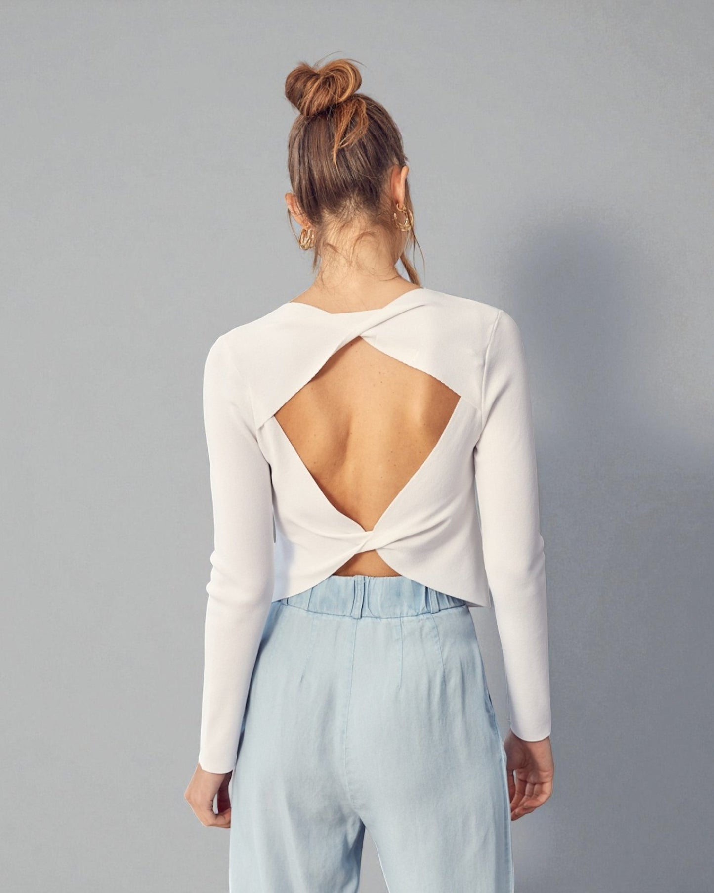 Person wearing a white long-sleeve top with an open back and light blue pants on a gray background