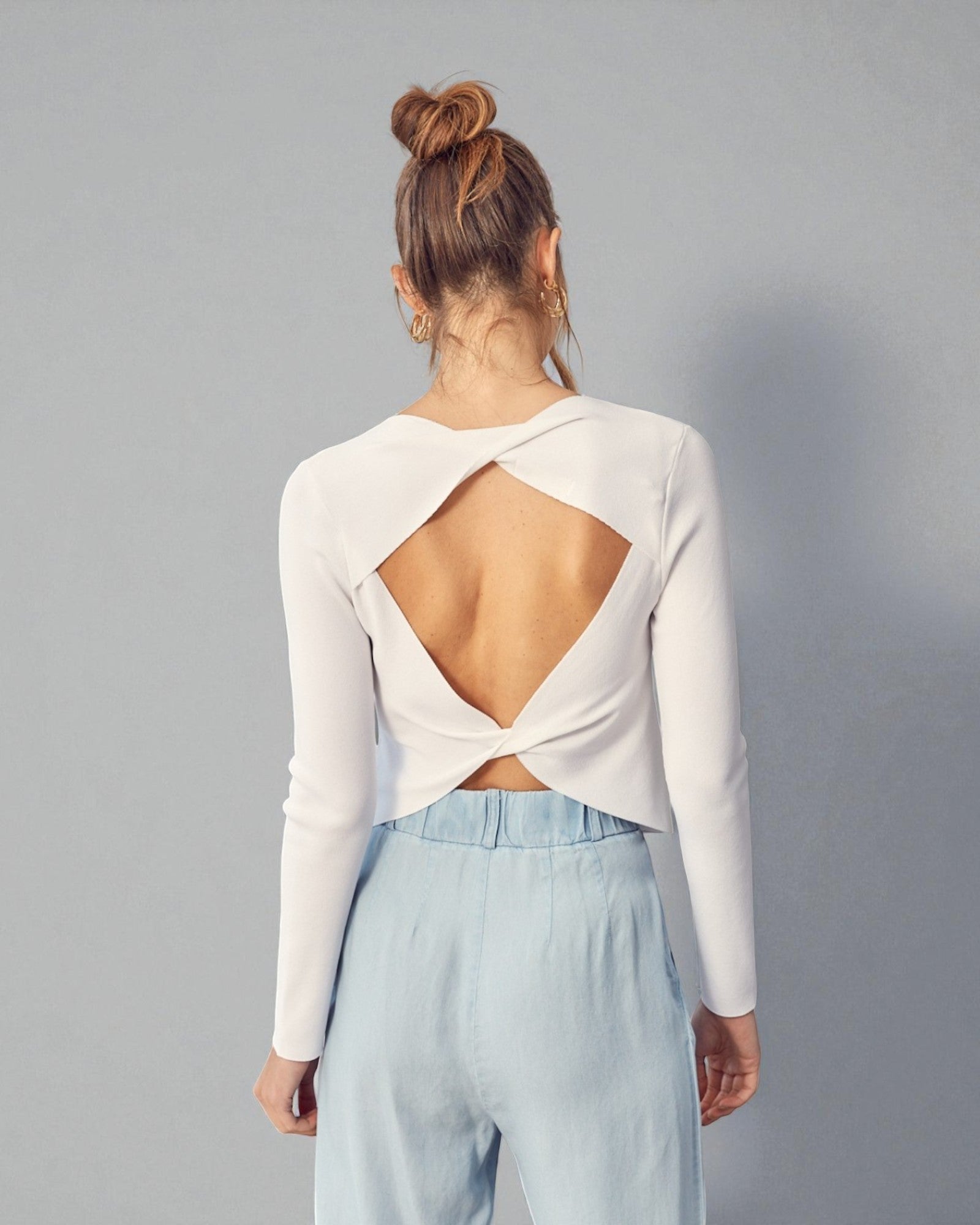 Person wearing a white long-sleeve top with an open back and light blue pants on a gray background
