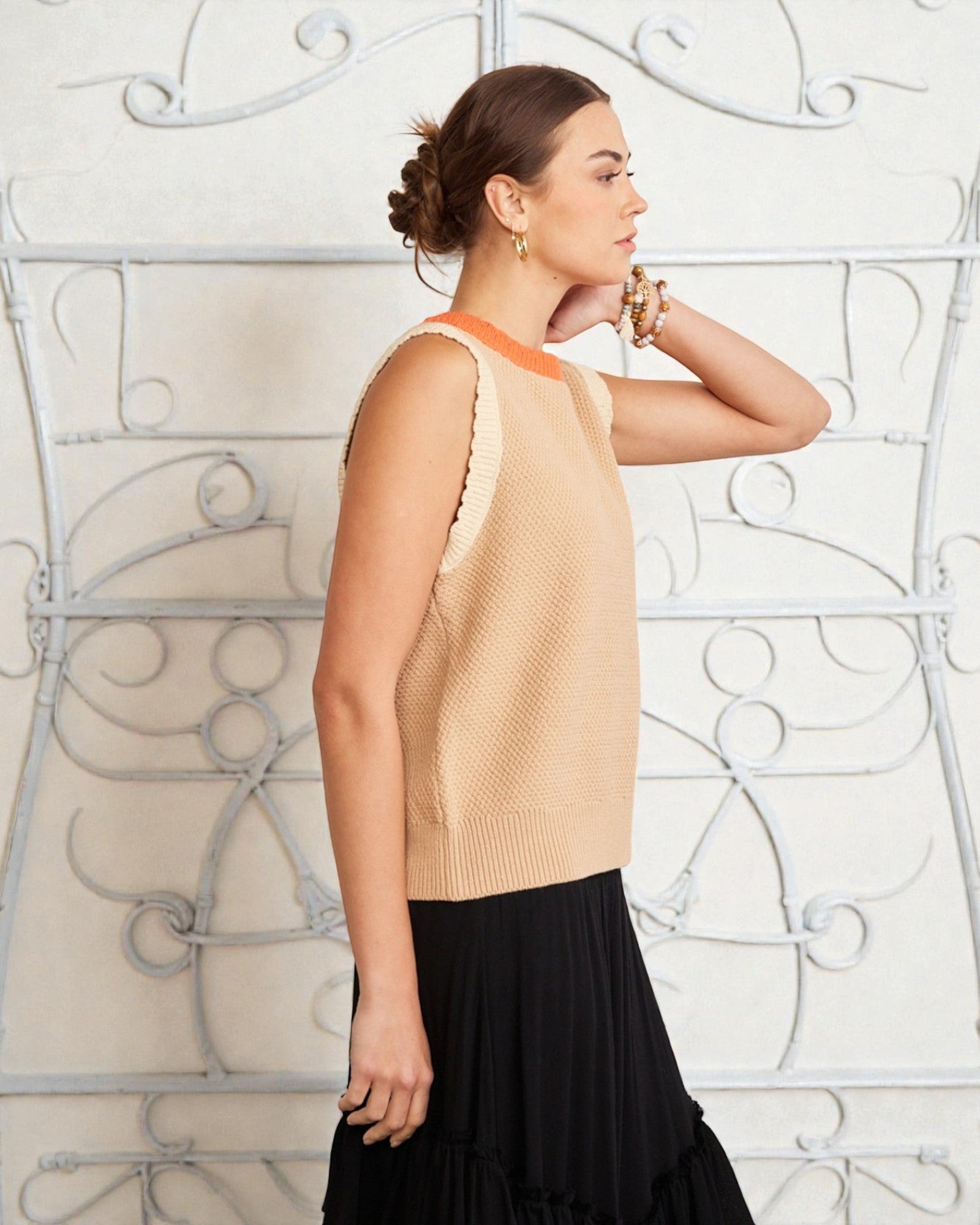 Woman wearing a beige sleeveless top with orange trim against an ornate white wall.