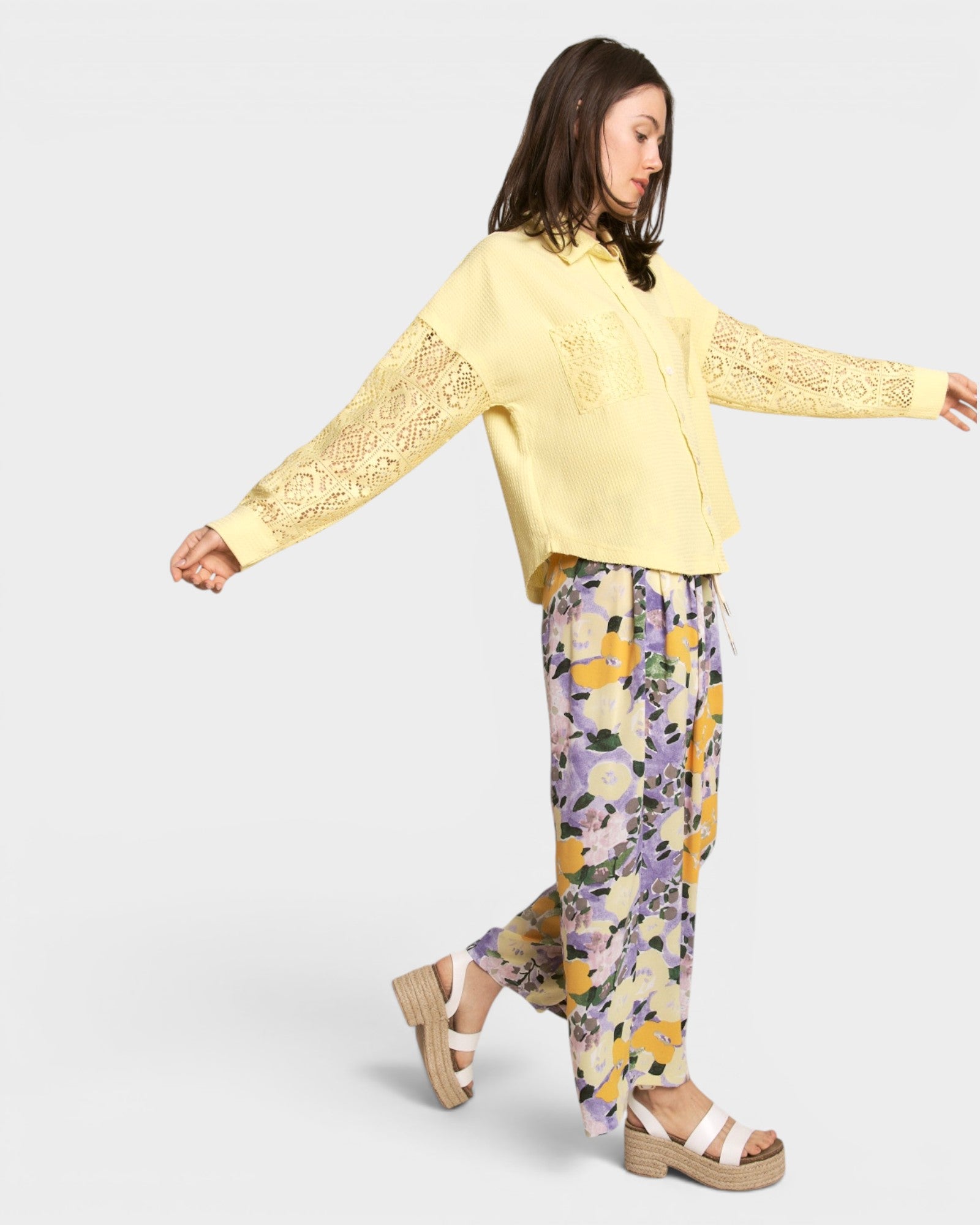 Woman wearing a yellow lace-trimmed top and floral pants on a light gray background