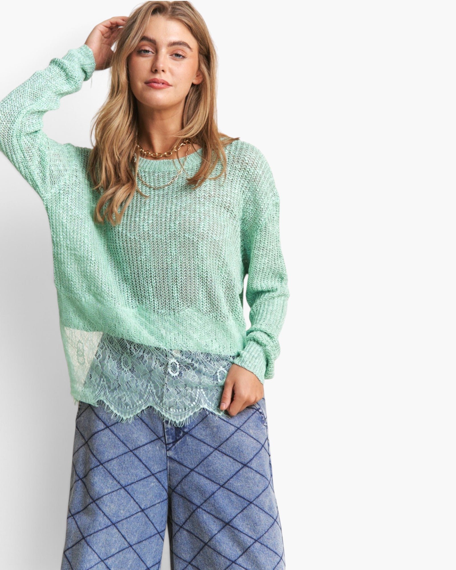 A woman wearing a light mint green textured knit sweater with a sheer lace hem, paired with blue argyle pants, against a plain white studio background.