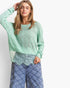 A woman wearing a light mint green textured knit sweater with a sheer lace hem, paired with blue argyle pants, against a plain white studio background.