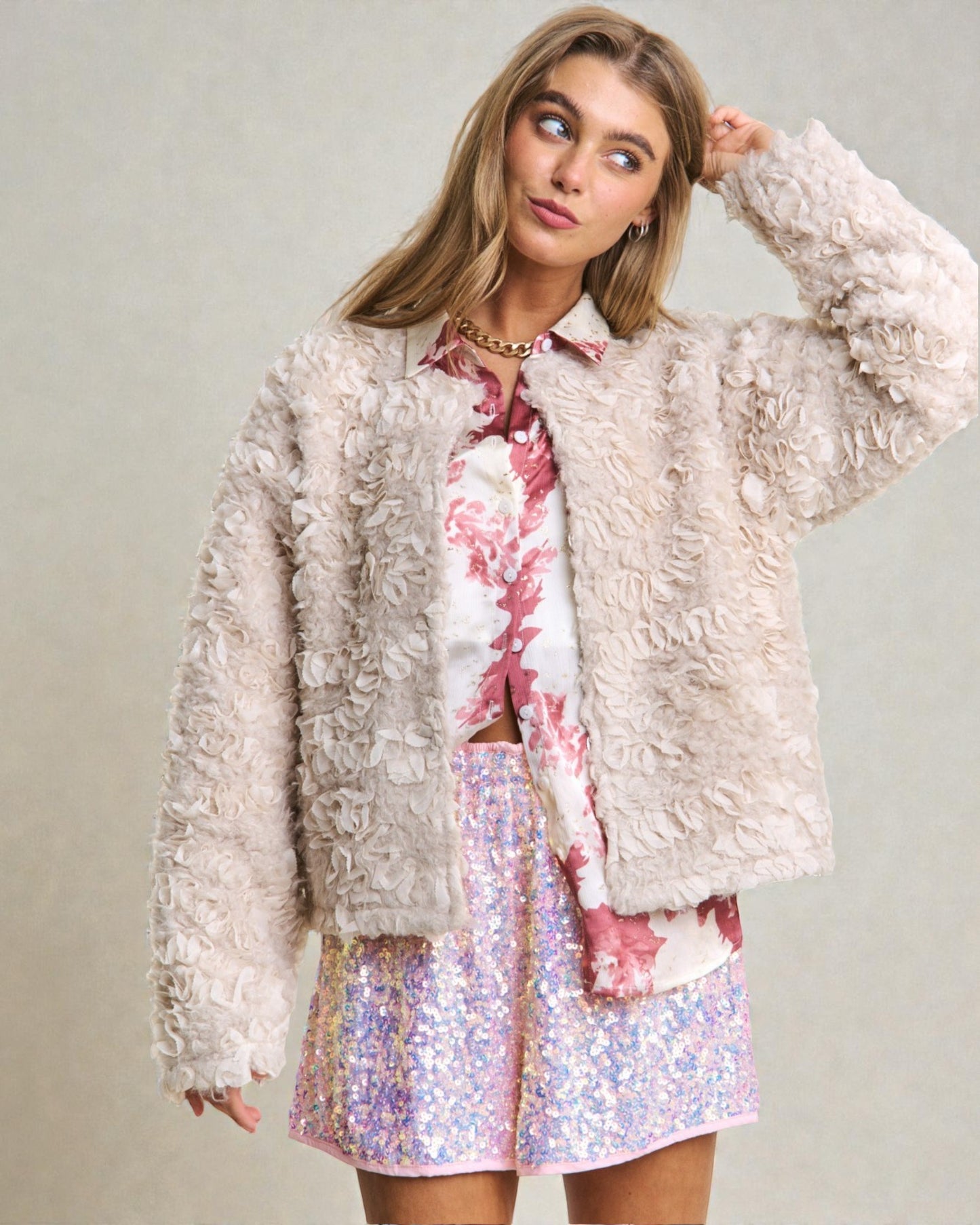 Woman wearing a textured beige jacket over a floral shirt and sequin skirt on a plain background