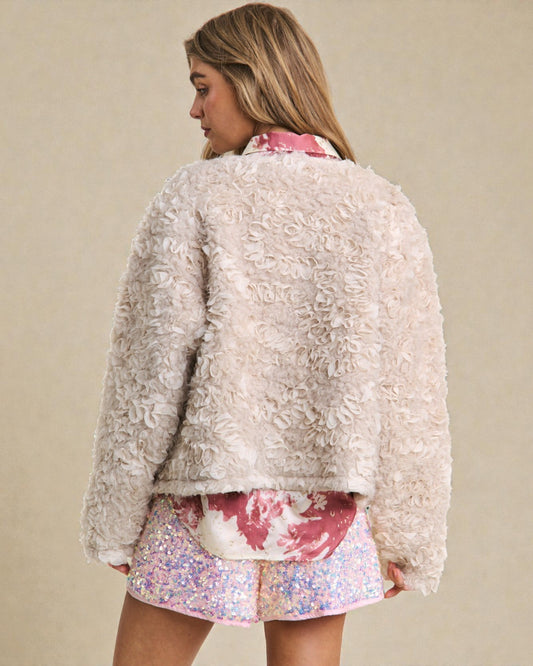 Woman wearing a textured beige jacket over a floral top and pink shorts on a beige background