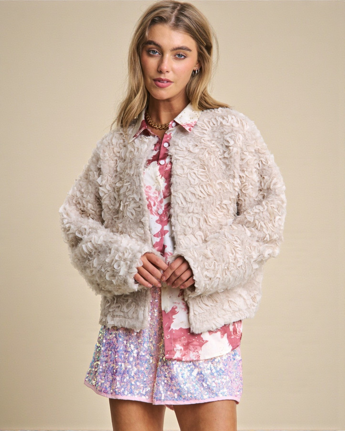 Woman wearing a textured beige jacket over a floral shirt and sequin skirt on a beige background