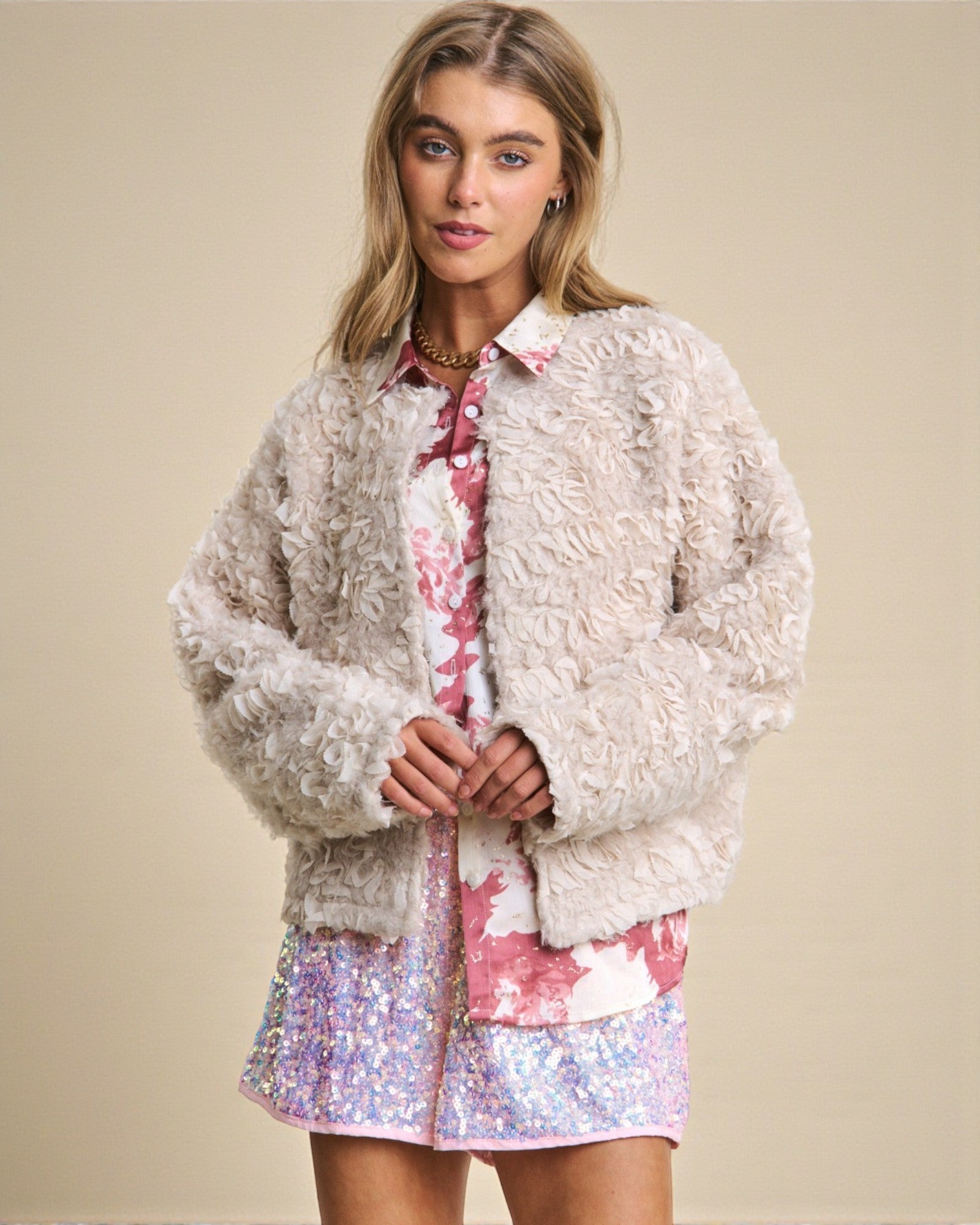 Woman wearing a textured beige jacket over a floral shirt and sequin skirt on a beige background