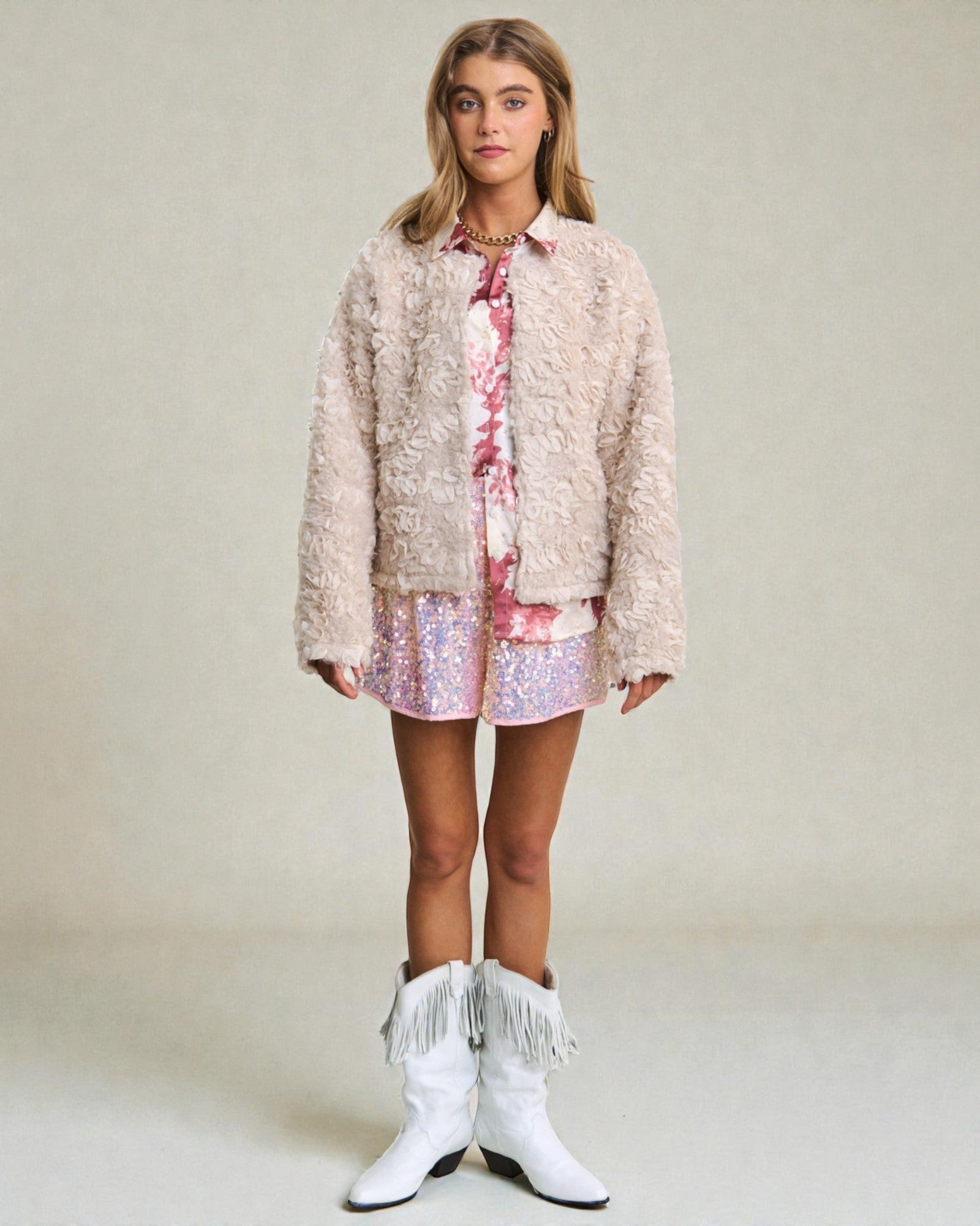 Woman wearing a fluffy beige jacket, floral shirt, sequin skirt, and white cowboy boots on a plain background