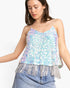 A woman wearing a light blue and silver iridescent sequin camisole top with fringe trim, paired with light-wash denim jeans, against a plain white studio background.