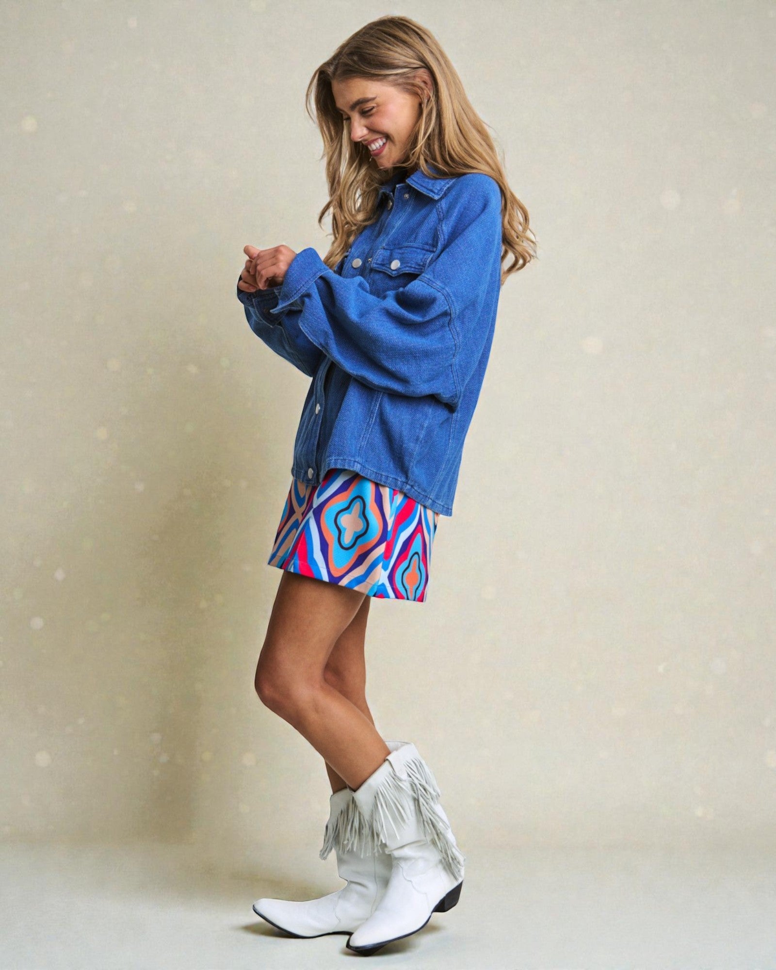 Woman wearing a blue denim jacket, colorful patterned skirt, and white boots on a beige background
