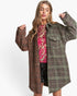 A woman modeling a long, oversized plaid coat in brown, green, and black tweed over a pink sequined top and black sequined shorts, set against a plain white studio background.