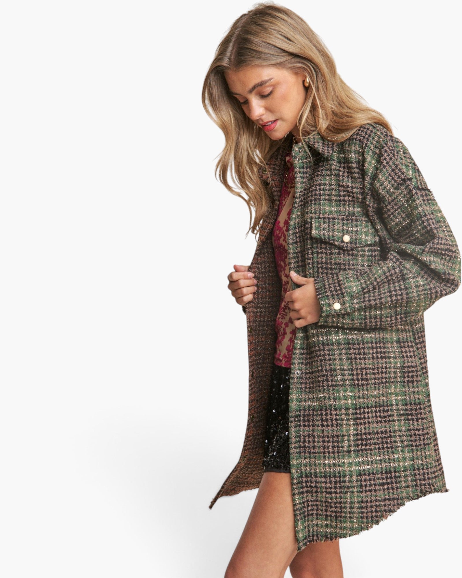 A woman wearing a long, wide-collar plaid coat in muted green, brown, and beige tones, paired with a patterned top and black shorts, photographed against a plain white studio background.