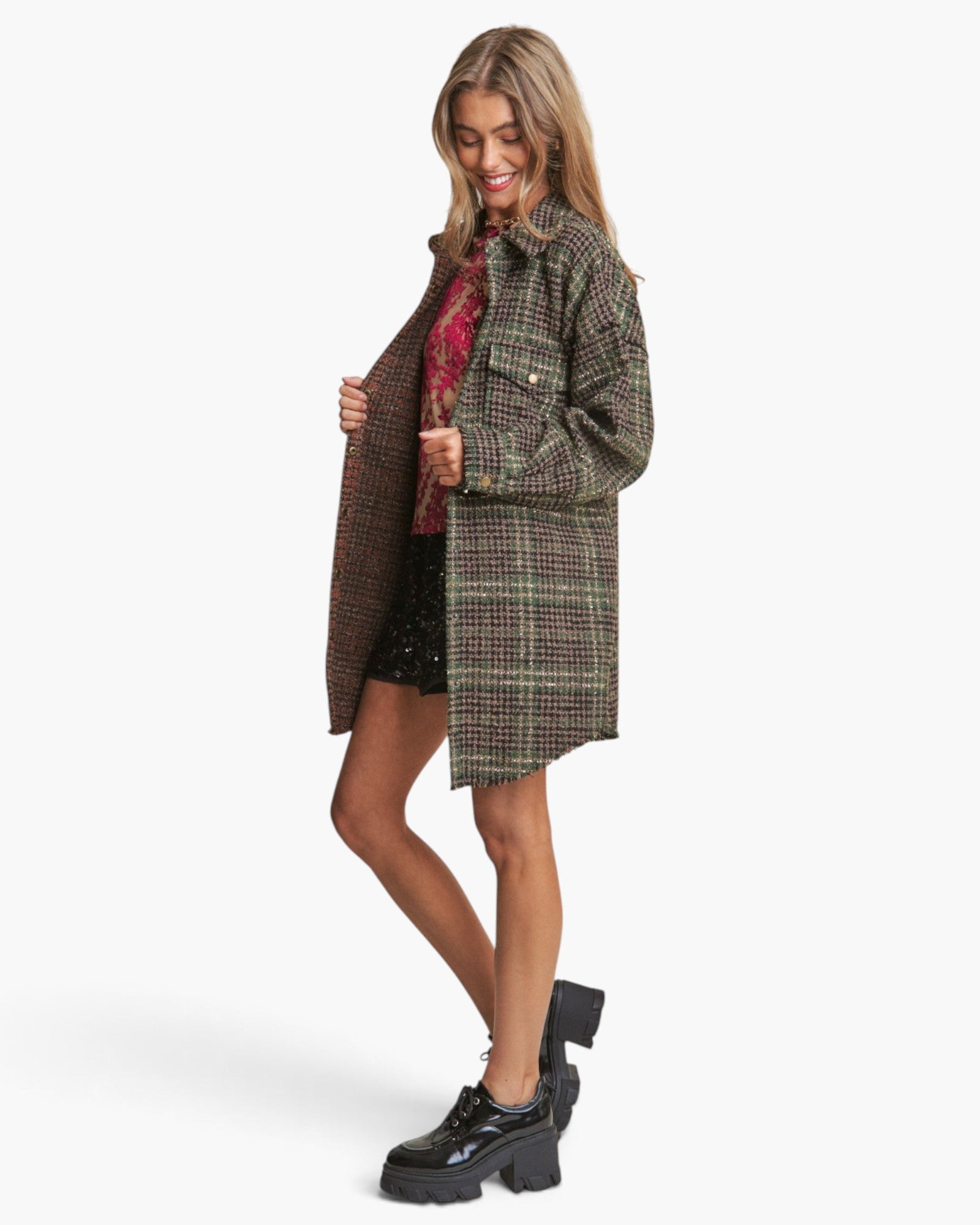 A woman modeling a brown and green plaid overshirt with a red patterned shirt underneath, black shorts, and black platform boots against a light gray background.