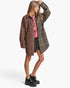 A woman modeling a brown and green plaid overshirt with a red patterned shirt underneath, black shorts, and black platform boots against a light gray background.