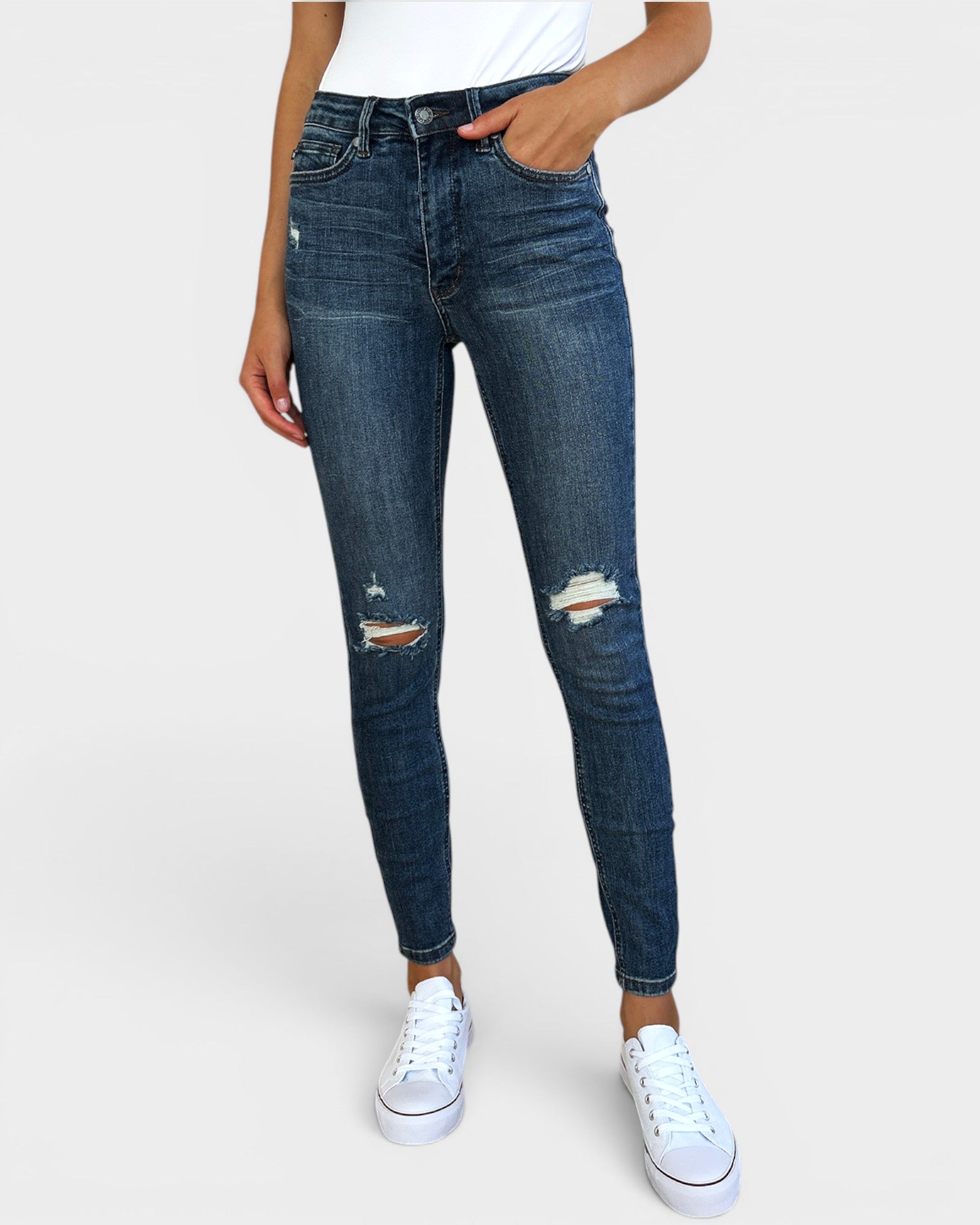 Person wearing blue jeans with slight distressing and white sneakers on a plain background