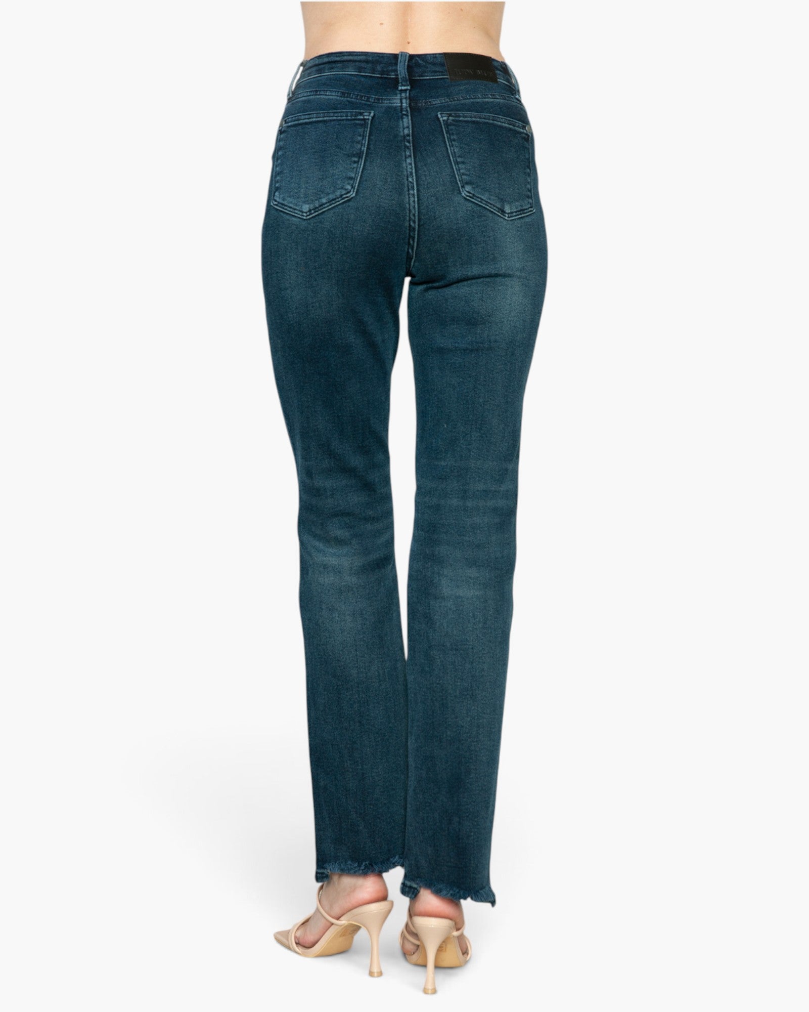 Rear view of a person wearing dark blue denim jeans with a straight-to-flare leg and frayed hems, paired with beige high-heeled sandals against a light gray studio background.
