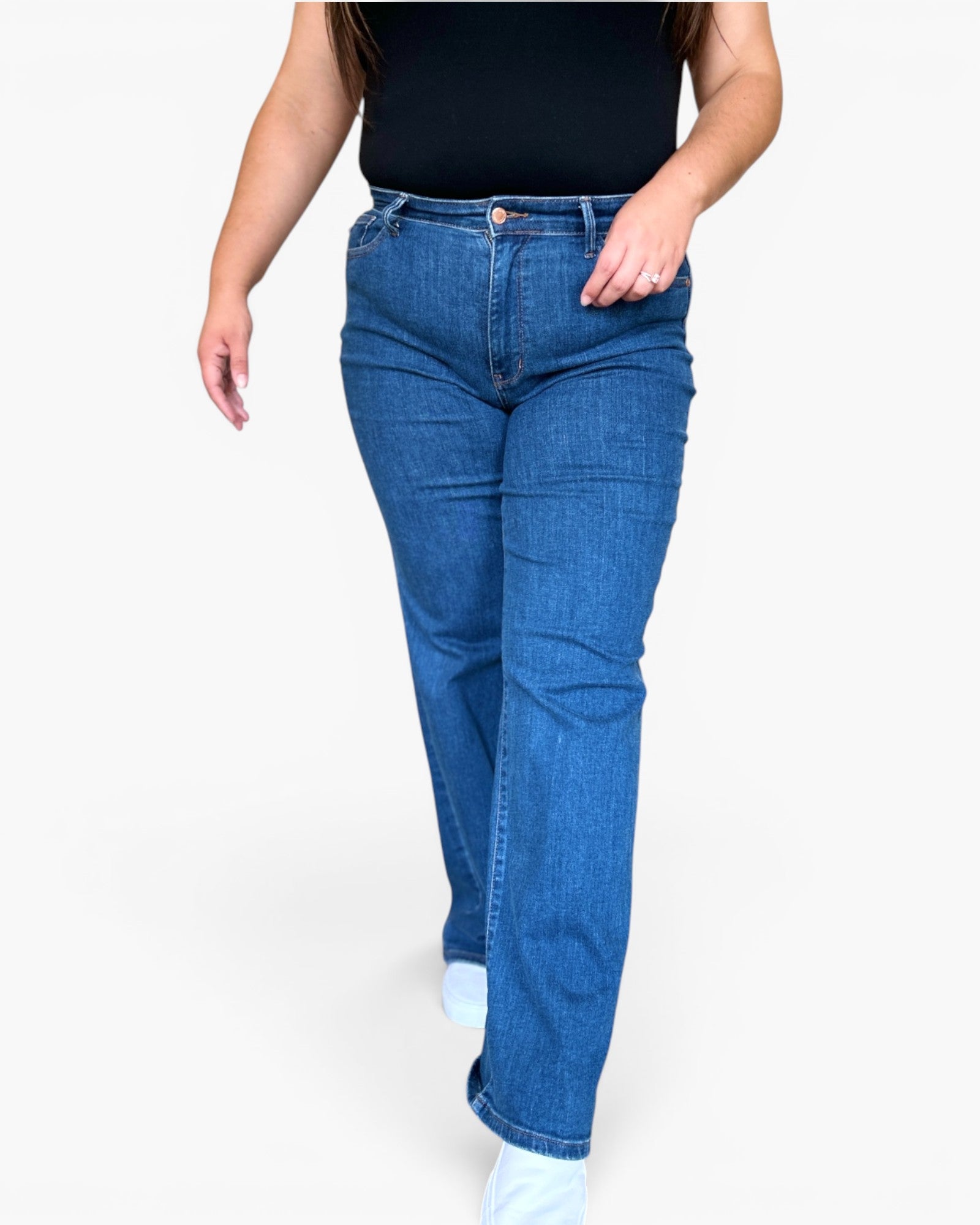 A person wearing medium-wash blue denim jeans with a black top and white socks, shown from the chest down against a plain light gray studio background.