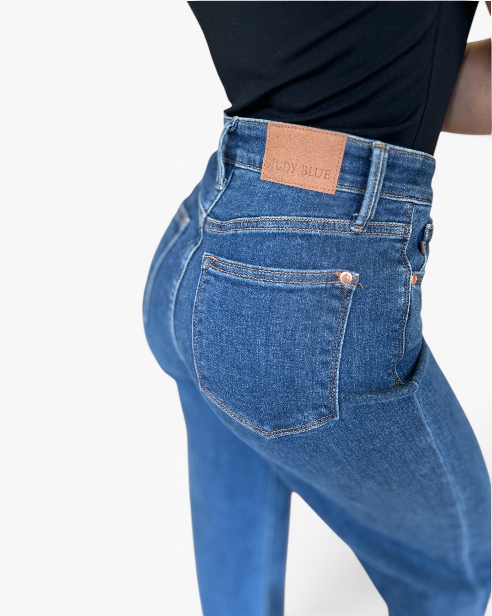 Close-up back view of a person wearing blue denim jeans with a black top, showing the waistband, belt loops, back pocket, and a tan leather patch that reads JUDY BLUE.