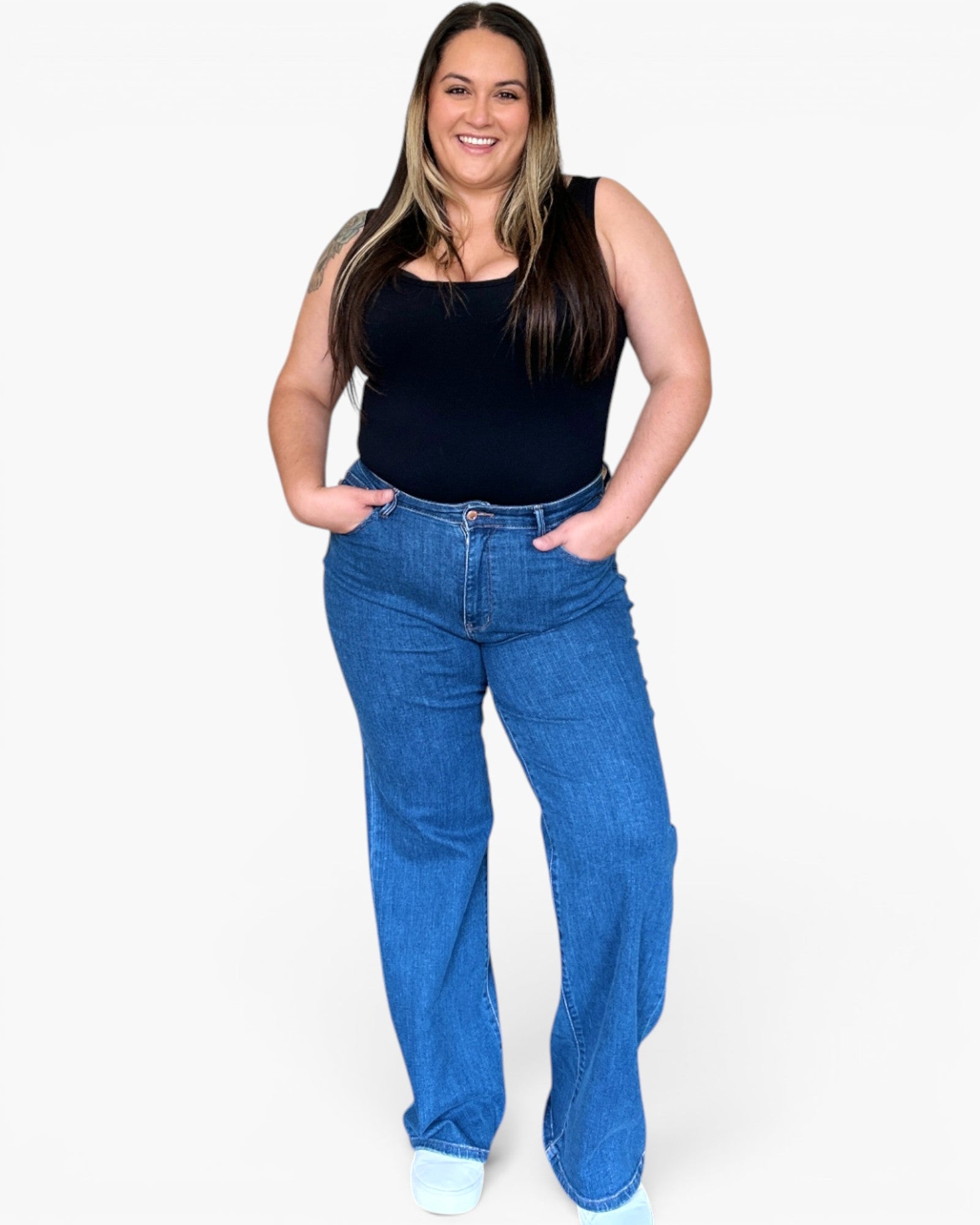 A woman modeling a pair of wide-leg blue jeans with a black sleeveless top, standing against a plain white background.