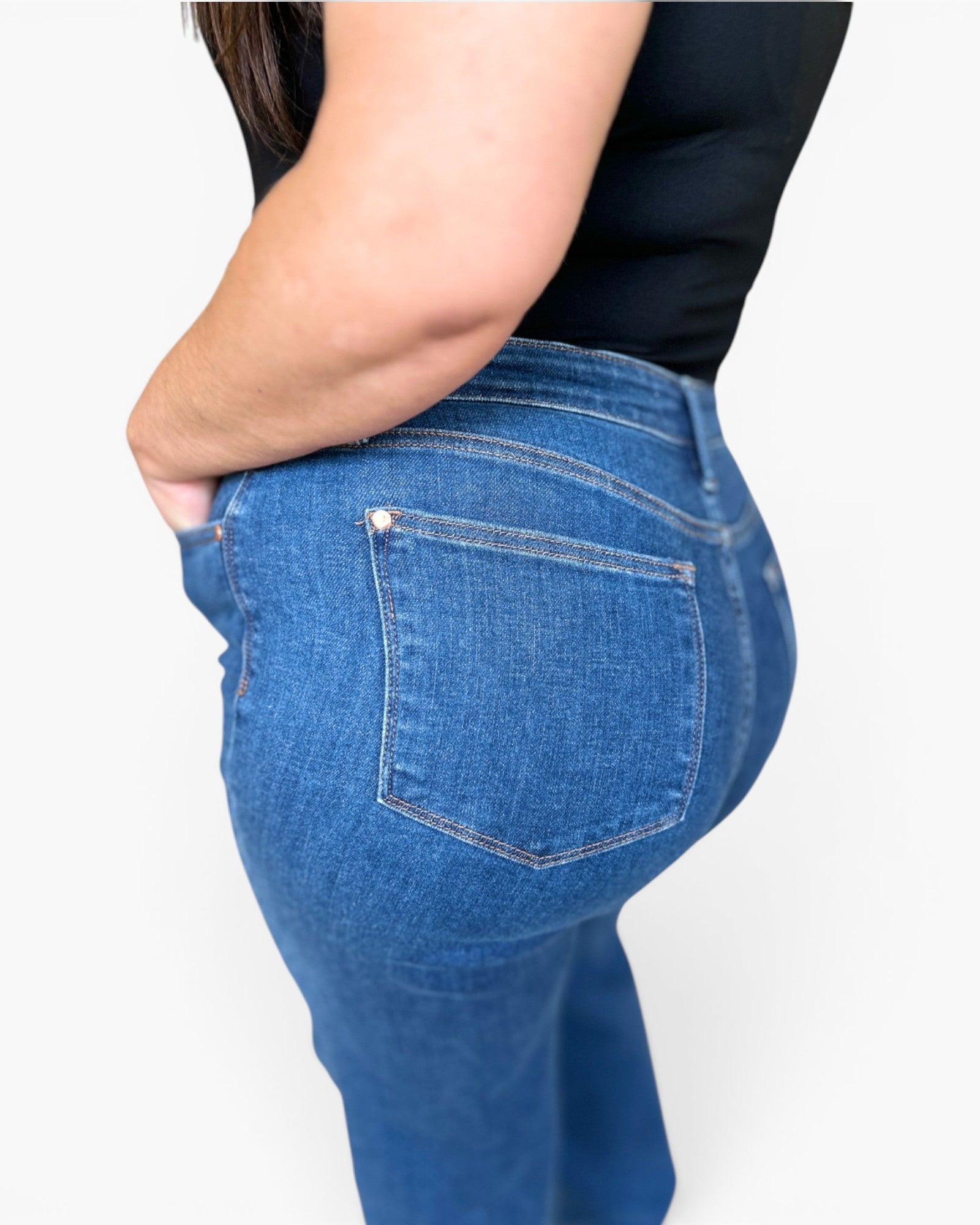 Close-up side and back view of a person wearing blue denim jeans with a black top against a light gray studio background.