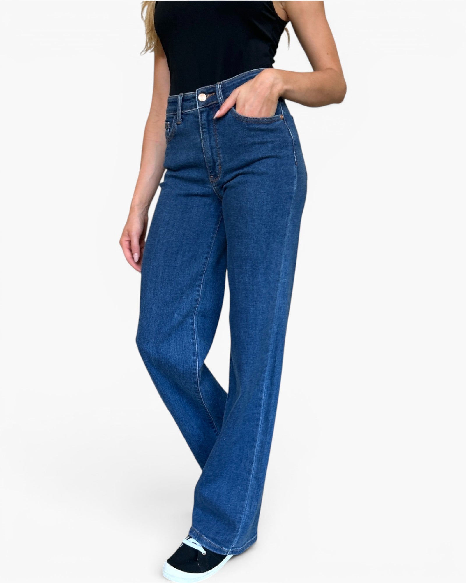 A model wearing a pair of high-waisted, wide-leg blue denim jeans with a black sleeveless top and black sneakers, shown from the waist down against a plain white background.
