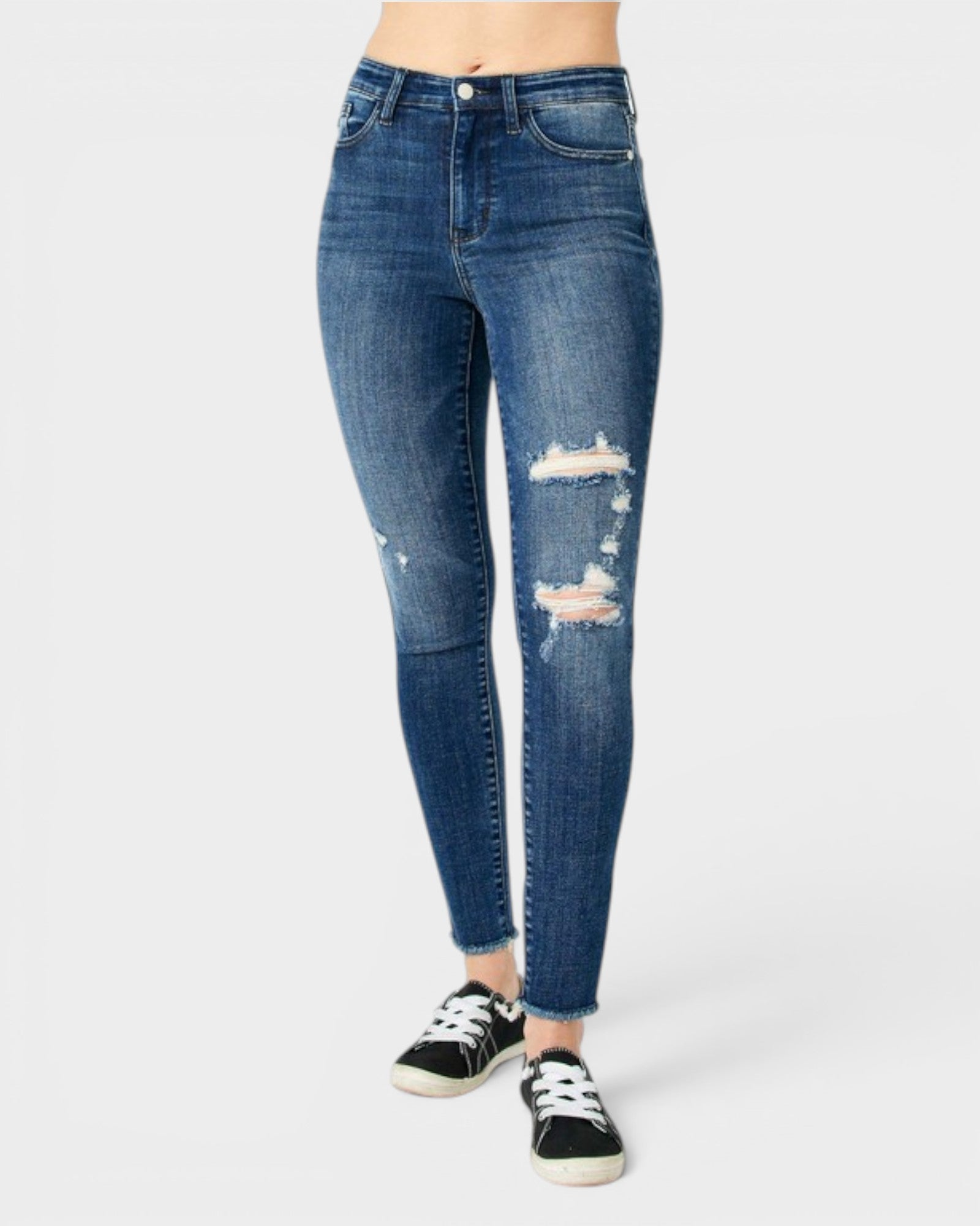 A person wearing blue skinny jeans with distressed rips on the thighs and knees, paired with black low-top sneakers, photographed against a light gray studio background.