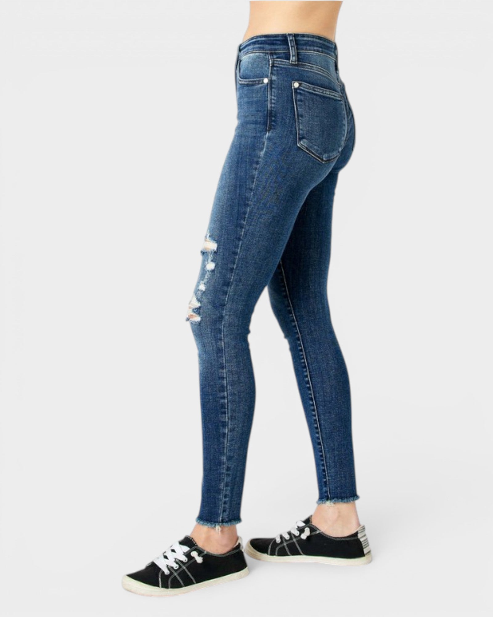 A side profile view of a person wearing dark blue distressed skinny jeans and black low-top sneakers against a light gray background.