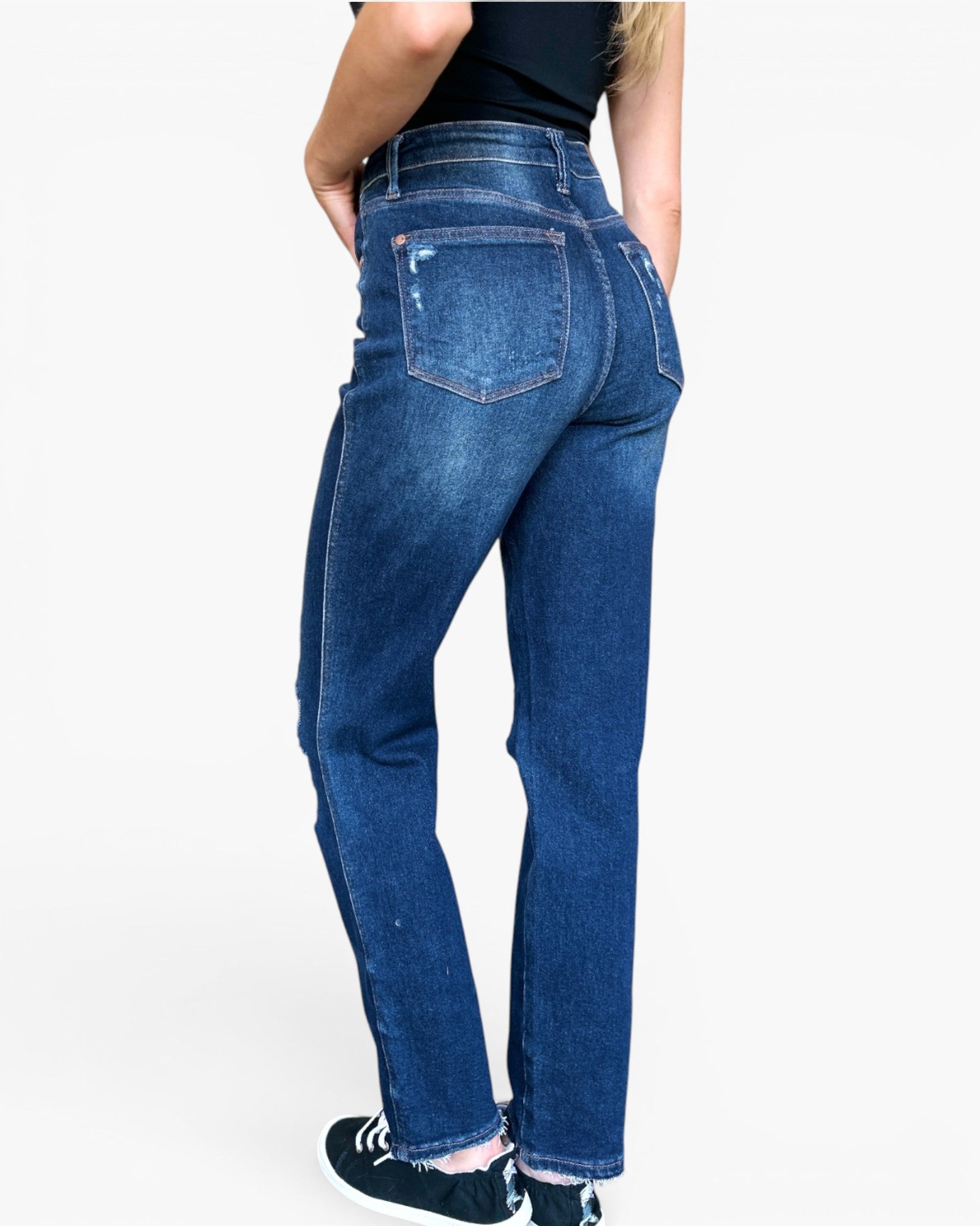 Rear view of a person wearing blue denim jeans with a black top and black sneakers against a light gray studio background.