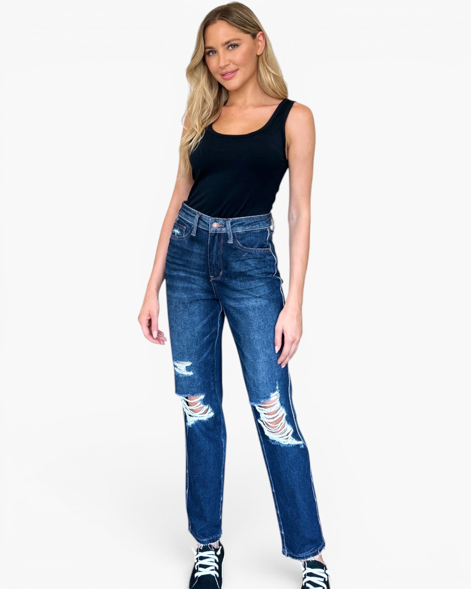 A woman modeling a pair of high-waisted, dark blue distressed jeans with ripped details on the thighs and knees, paired with a black tank top and black sneakers, against a plain white background.