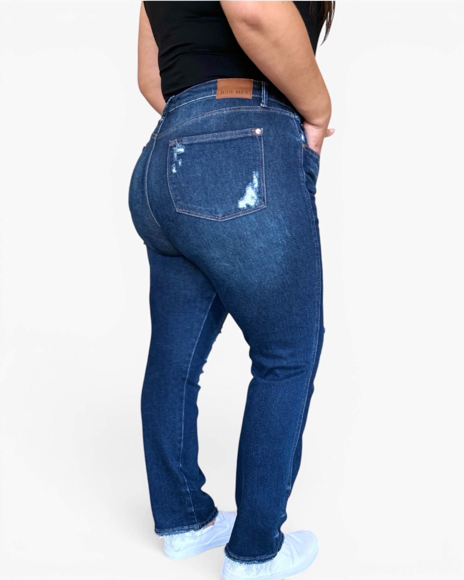 A person shown from the back wearing dark blue distressed jeans with a black top and white sneakers, photographed against a plain light background.