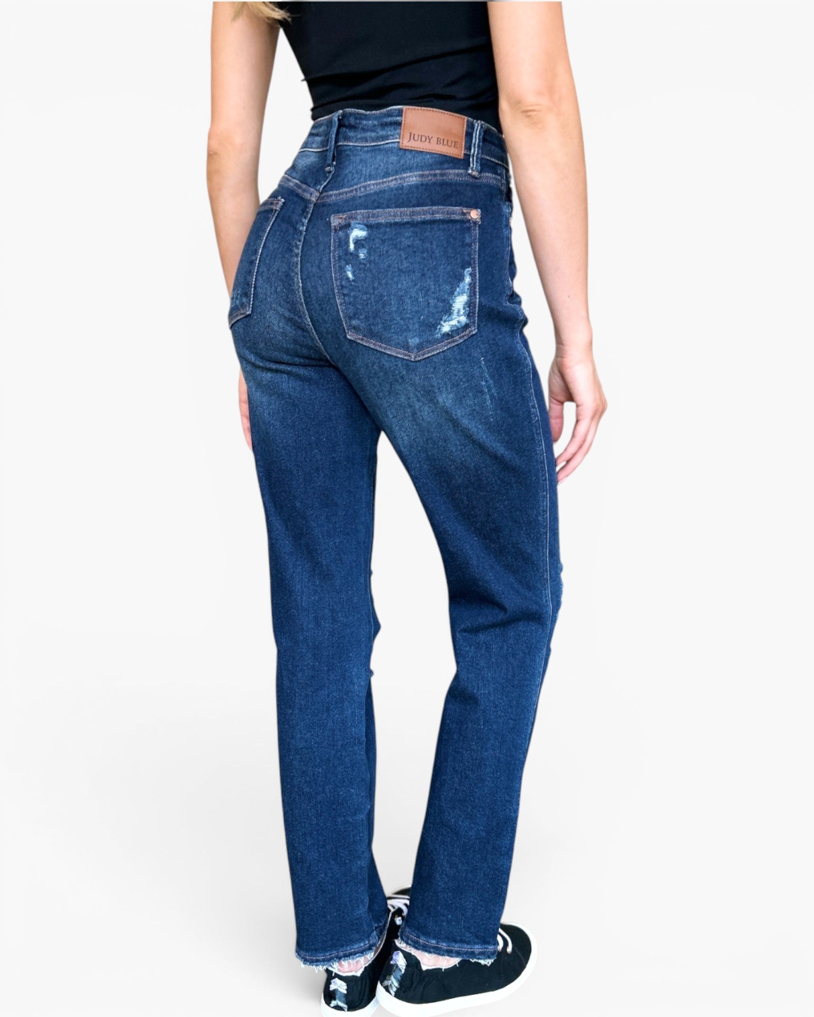 A person shown from the back wearing dark blue high-waisted jeans with a distressed front pocket and a brown leather brand patch. The background is a clean white studio backdrop.