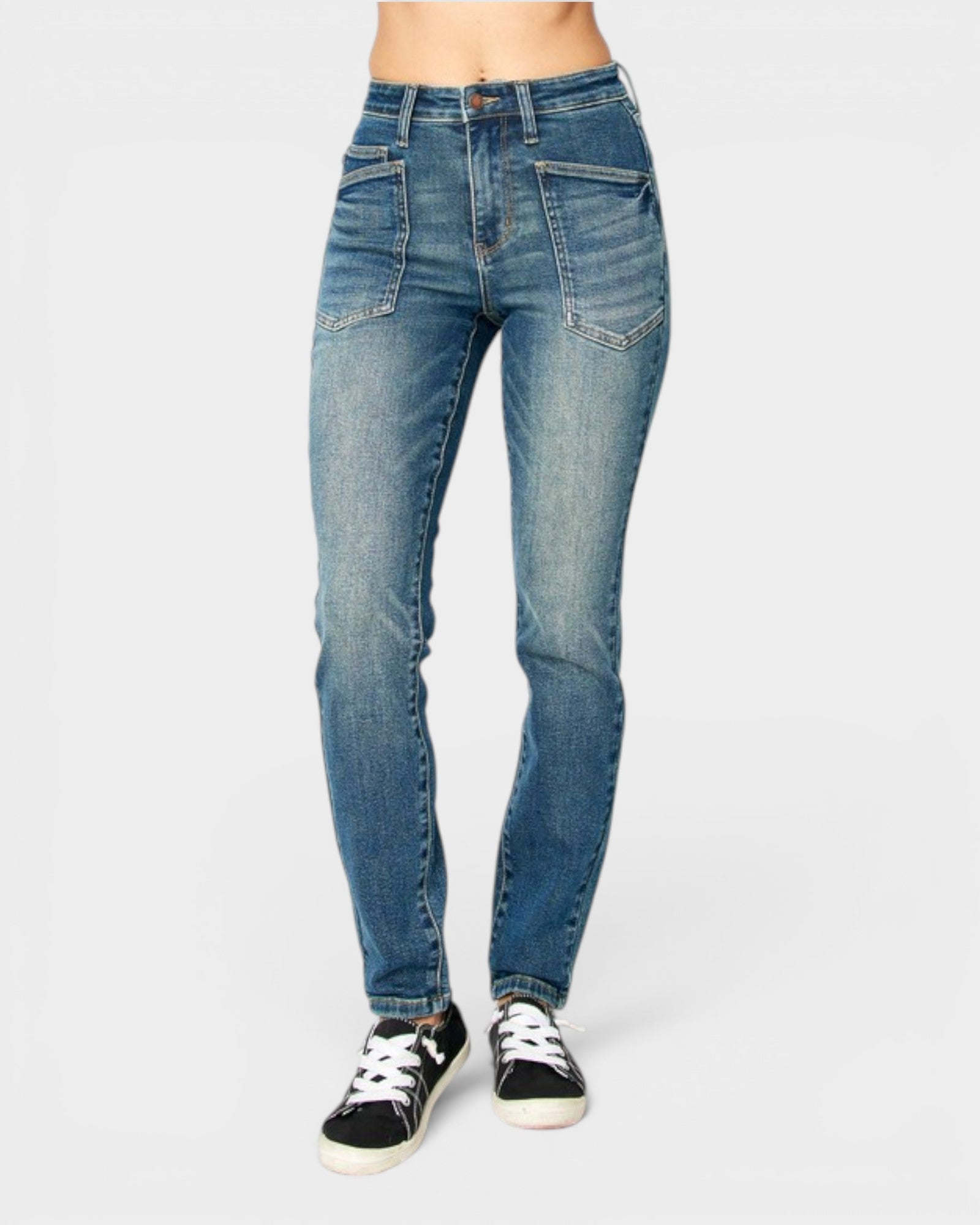 A person wearing medium-wash blue skinny jeans with black low-top sneakers, shown from the waist down against a light gray studio background.