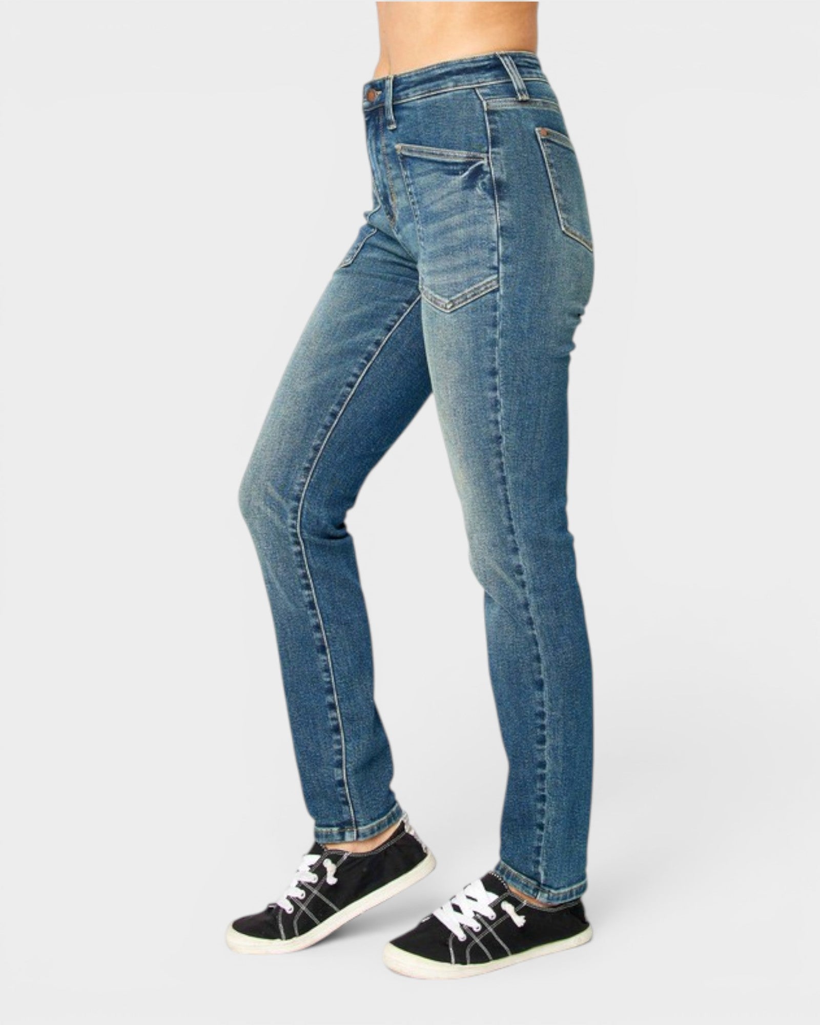 A person wearing medium-wash blue skinny jeans and black canvas sneakers, shown from the waist down against a light gray studio background.